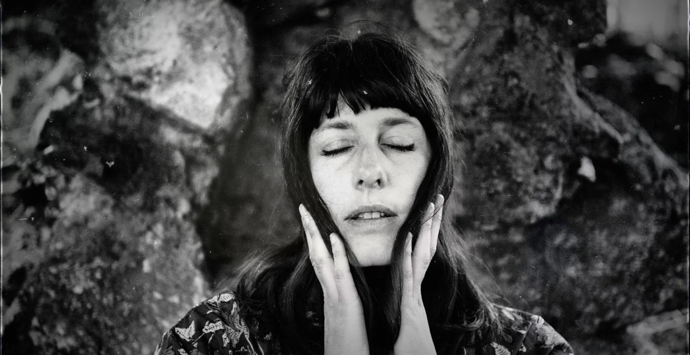 A black and white photo of a woman with her eyes closed, holding her face with both hands, standing outdoors with trees in the background.