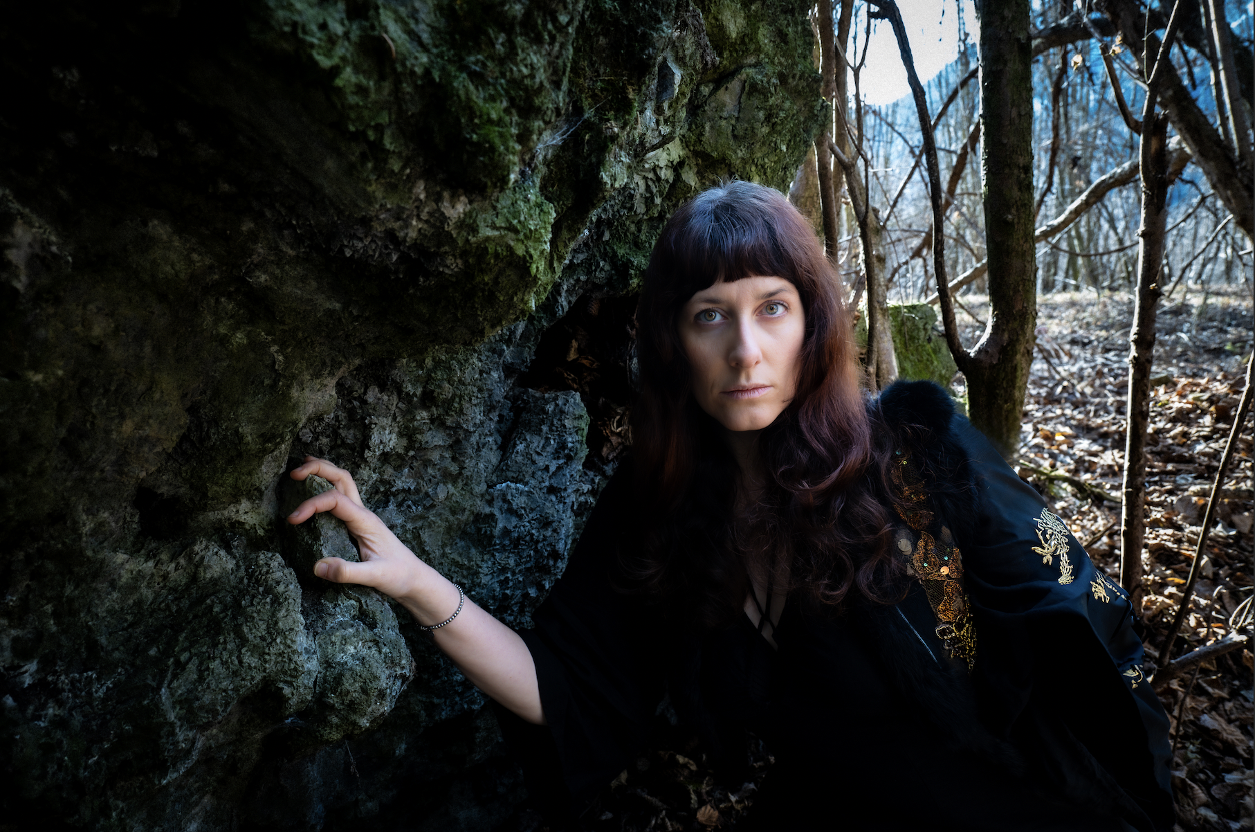 A woman with long dark wavy hair and blue eyes crouches near a mossy rock in a forest, with trees and fallen leaves in the background.