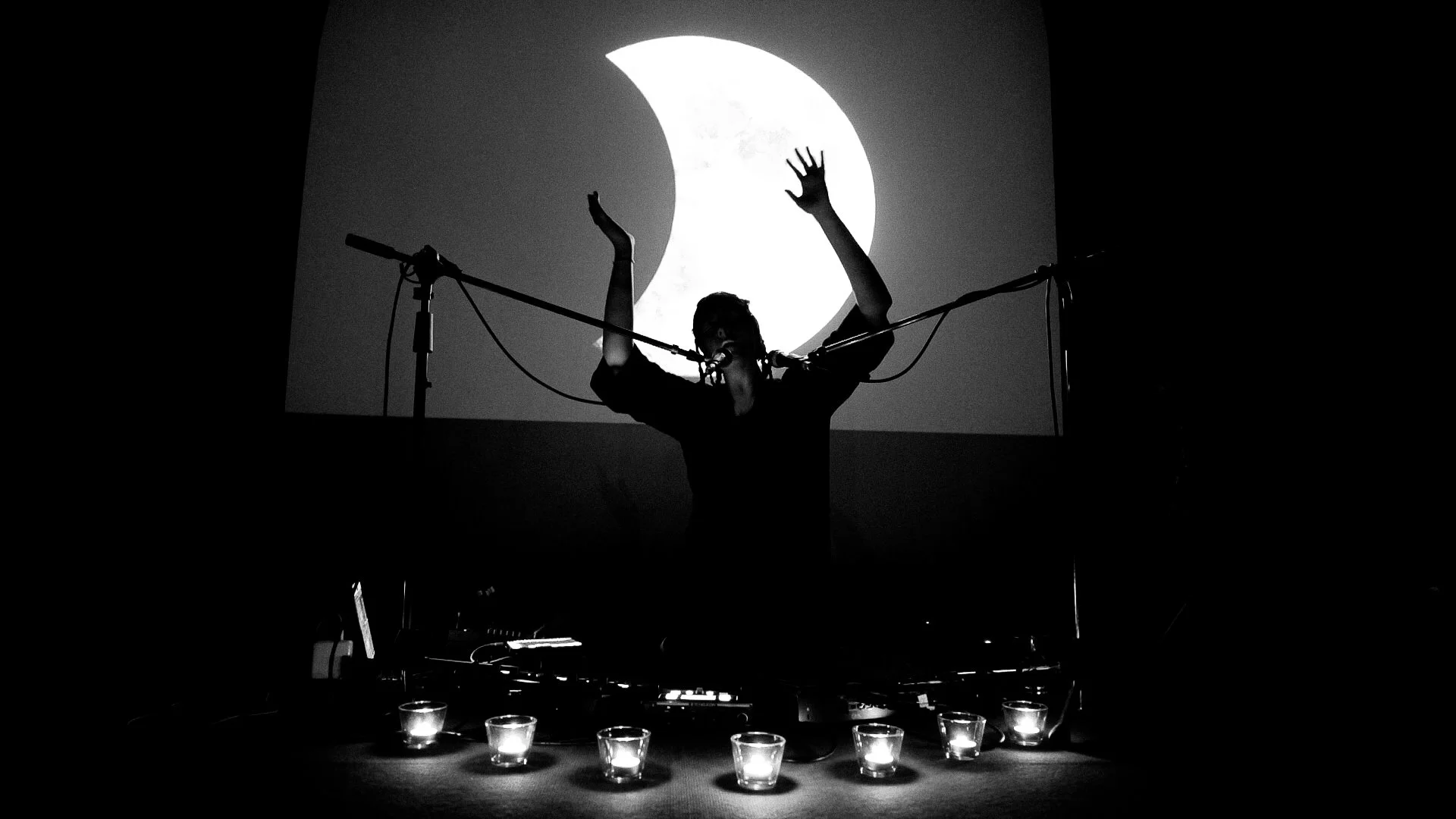 A person performing on stage with arms raised, standing in front of a large projected image of the moon, with candles arranged in front of them.