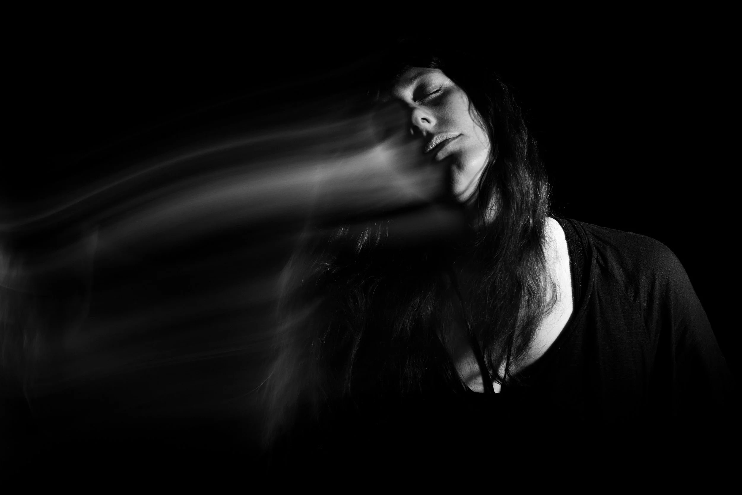 Black and white photo of a woman with long hair, her face tilted back and eyes closed, with a motion blur suggesting movement or wind, against a dark background.