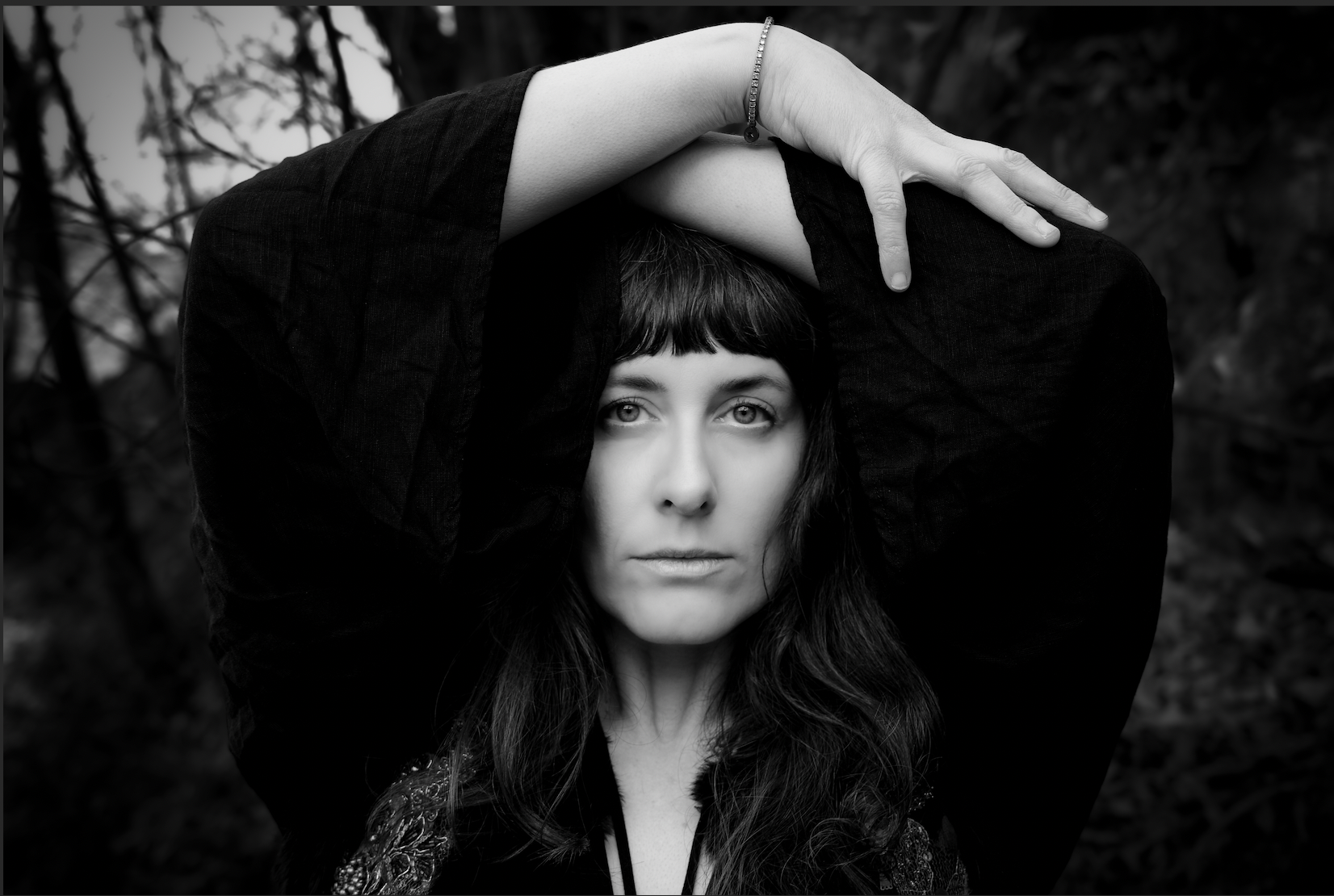 A woman with dark hair and bangs looking directly at the camera, holding her arms above her head, with a forest background in black and white.