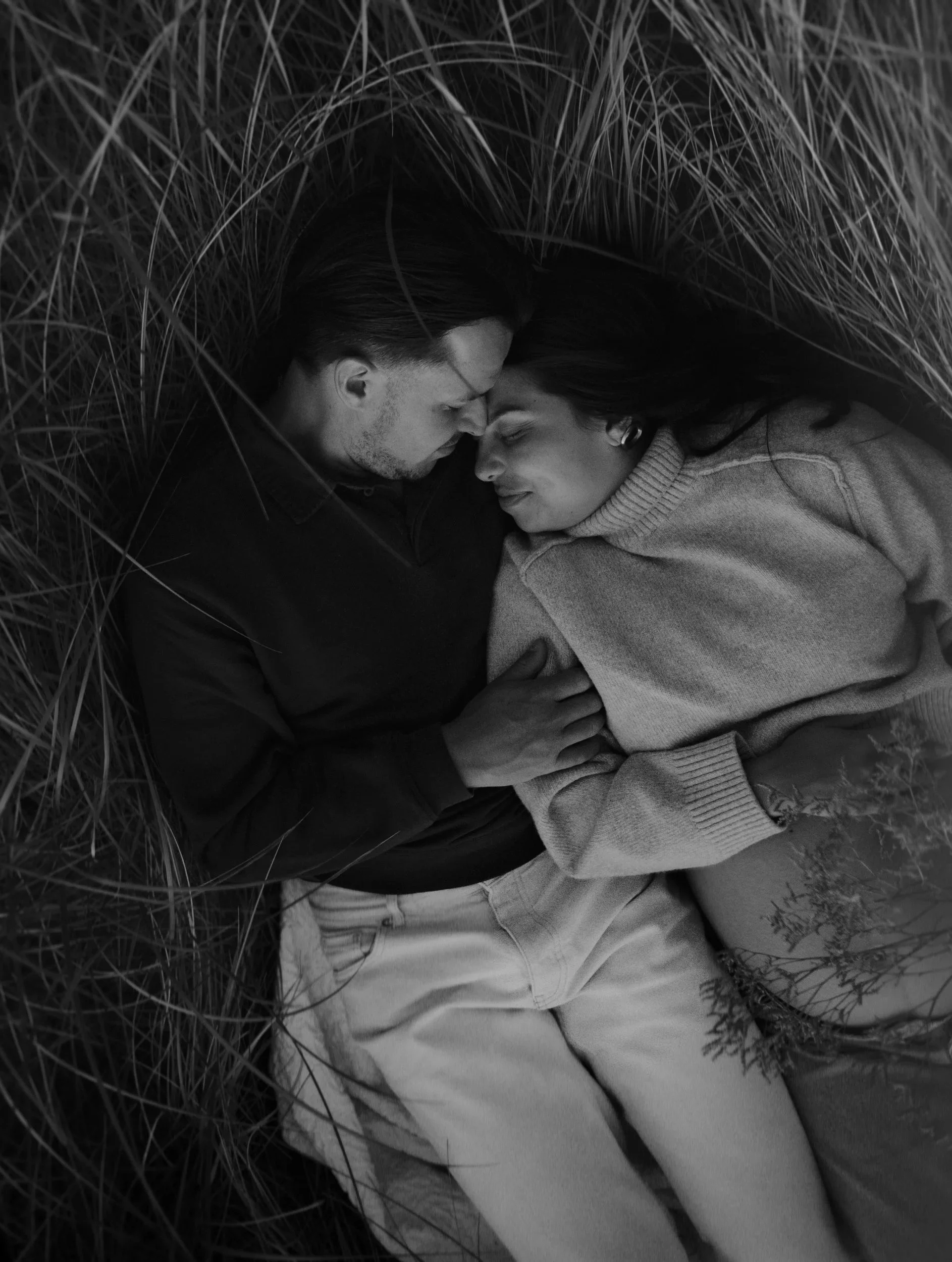 A black and white photo of a man and woman lying on the ground amidst tall grass, nose to nose, in a tender moment.