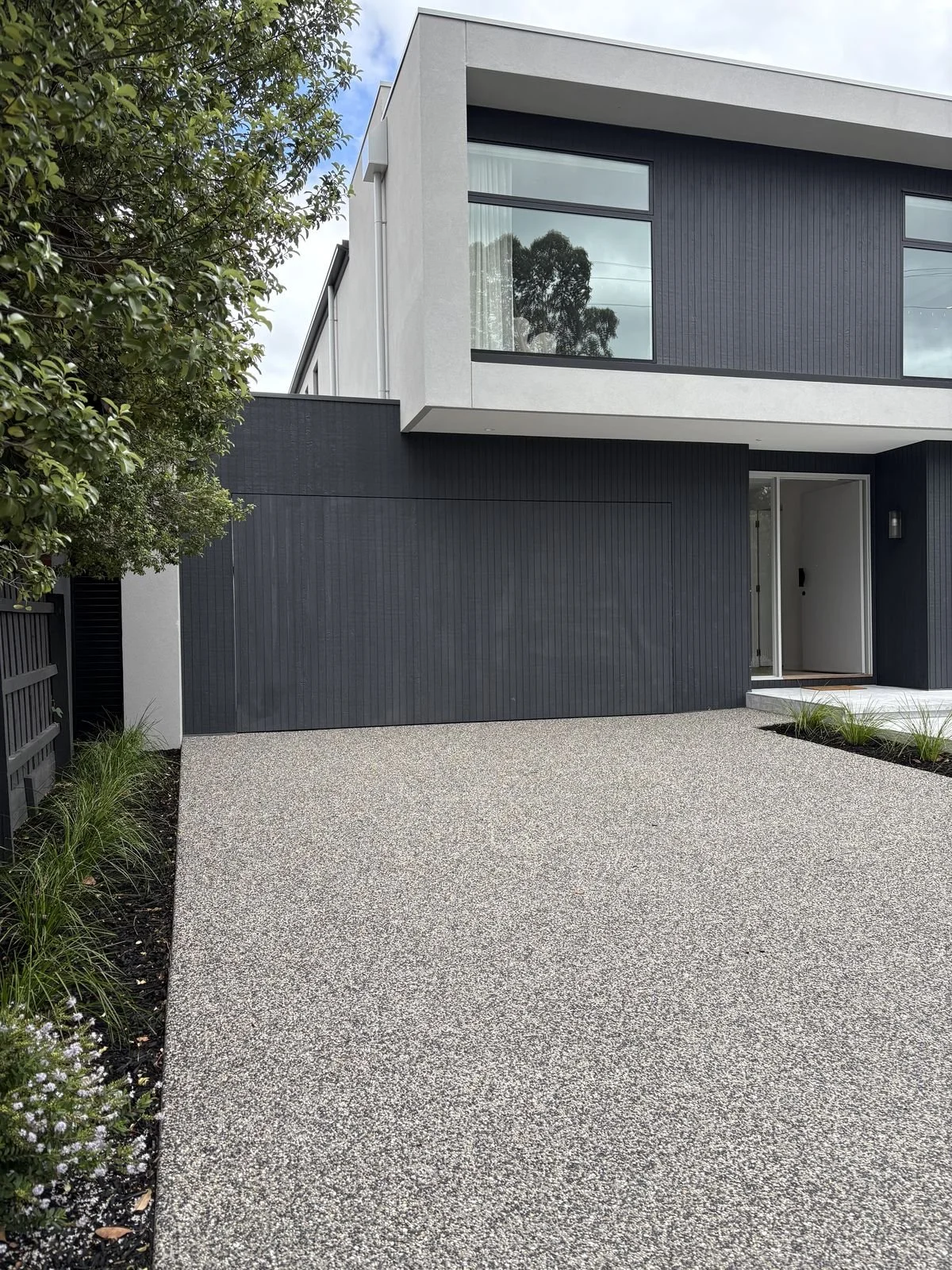 Modern two-story house with a gray exterior, large windows, a gravel driveway, and minimal landscaping.