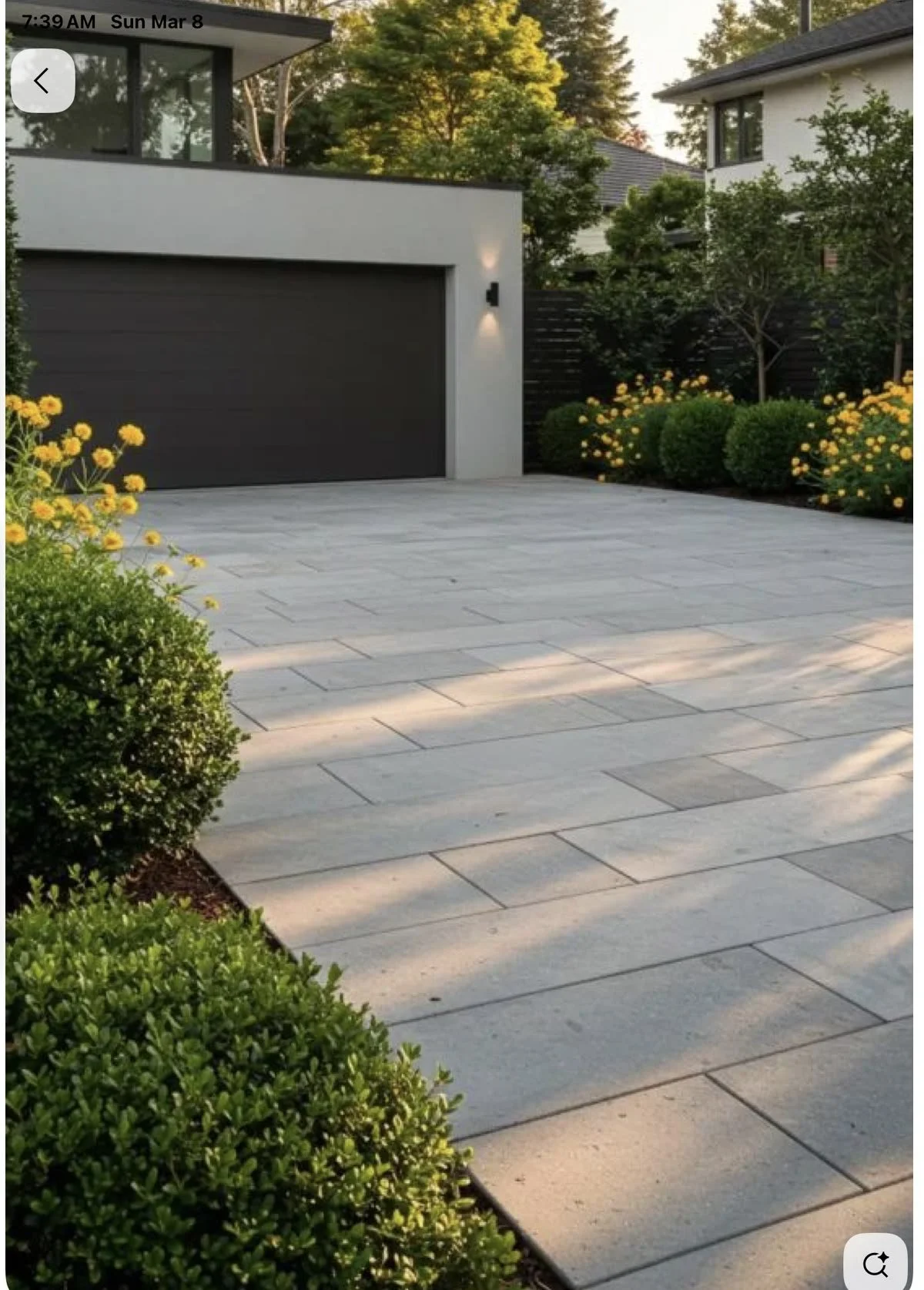 Modern house exterior with a dark gray garage door, surrounded by well-maintained green bushes and bright yellow flowers, and trees in the background. The driveway is paved with large light-colored stones, and there is an outdoor wall-mounted light fixture to the right of the garage door.