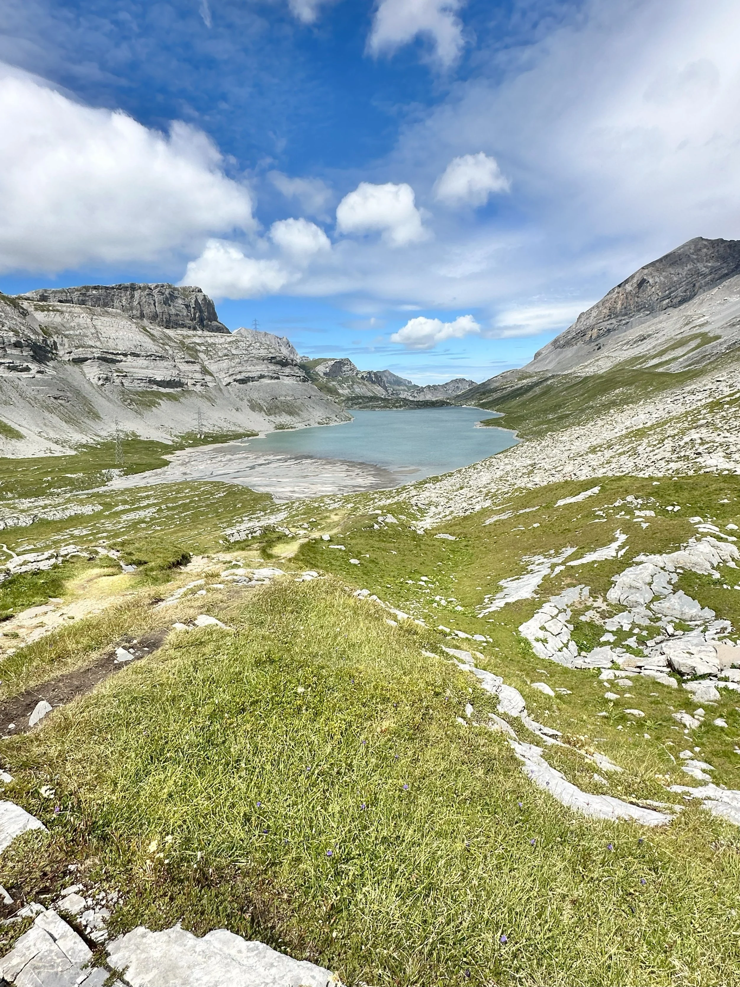 Bergsee in einer Alpenlandschaft mit grünem Gras, steinigen Ufern und hohen Bergen im Hintergrund, unter einem blauen Himmel mit Wolken.