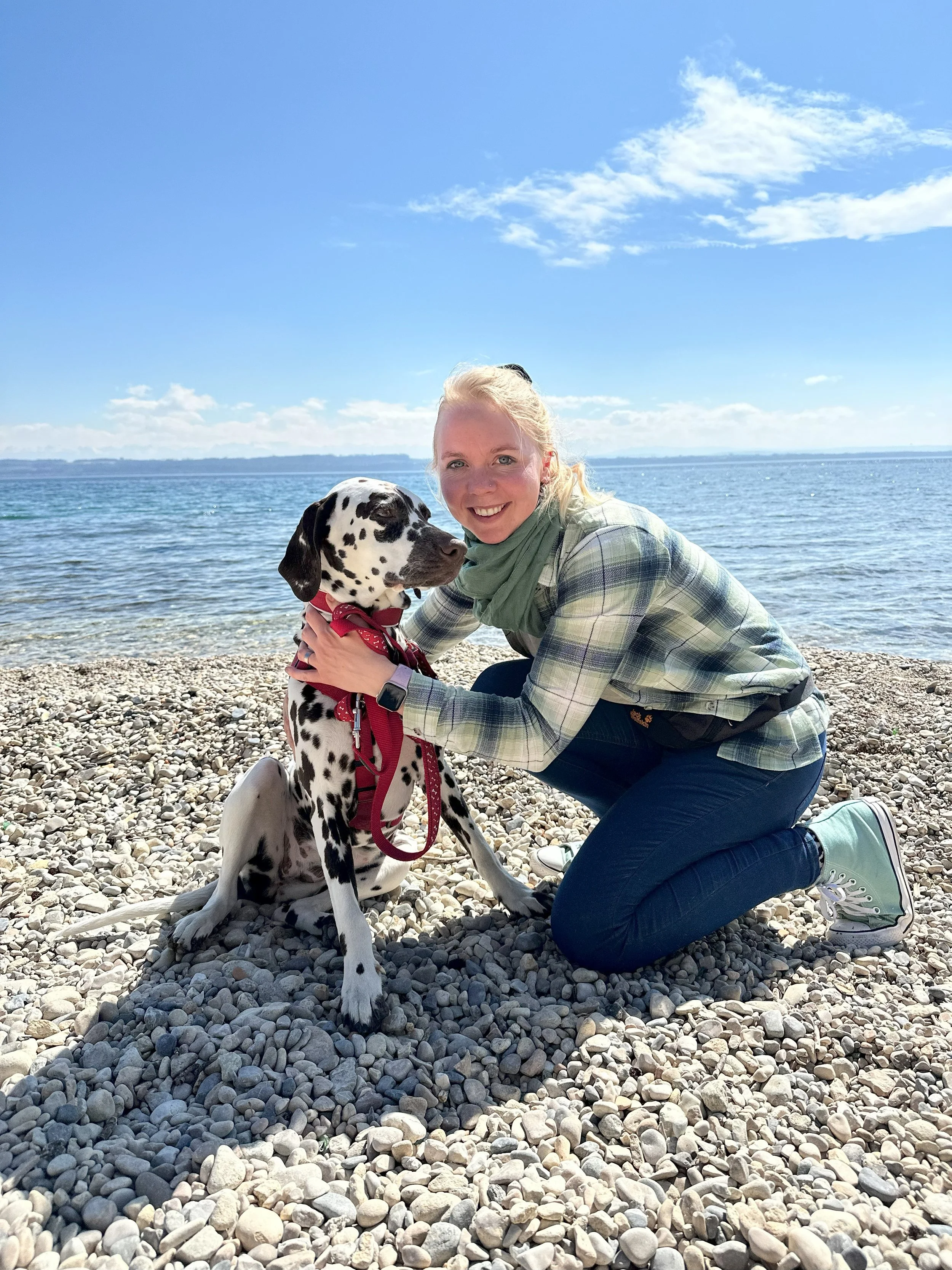 Eine lachende Frau kniet an einem Kieselstrand neben einem Dalmatiner-Hund mit rotem Geschirr und Leine, im Hintergrund ist das Meer und ein blauer Himmel mit Wolken.