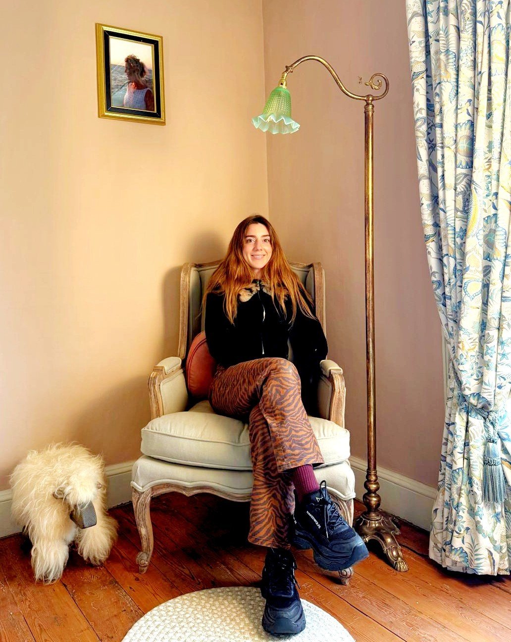 A woman with long hair sitting in a vintage armchair near a window with floral curtains, a standing floor lamp, a framed photo on the wall, and a fluffy white sheep sitting on the wooden floor next to her.