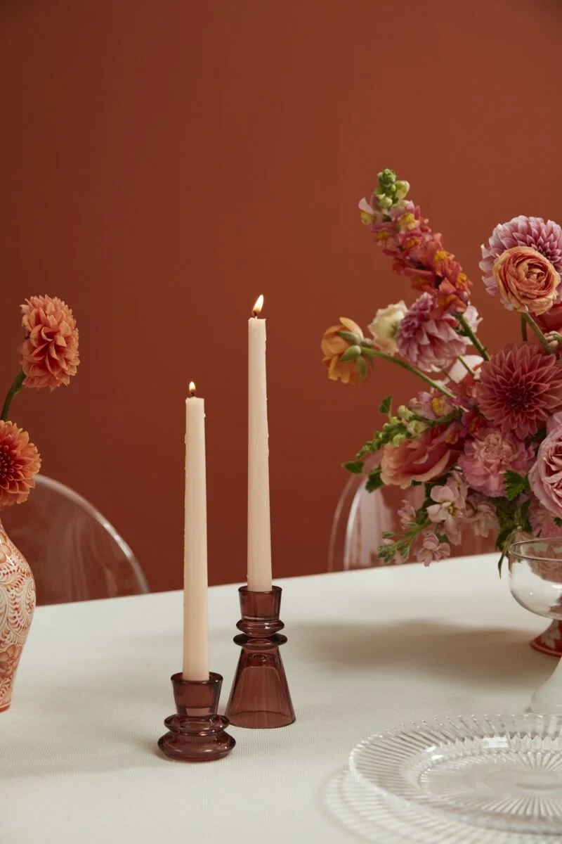 A table decorated with pink and peach flowers in vases, two lit white candles in amber glass candle holders, and clear glass plates, set against a warm brown background.