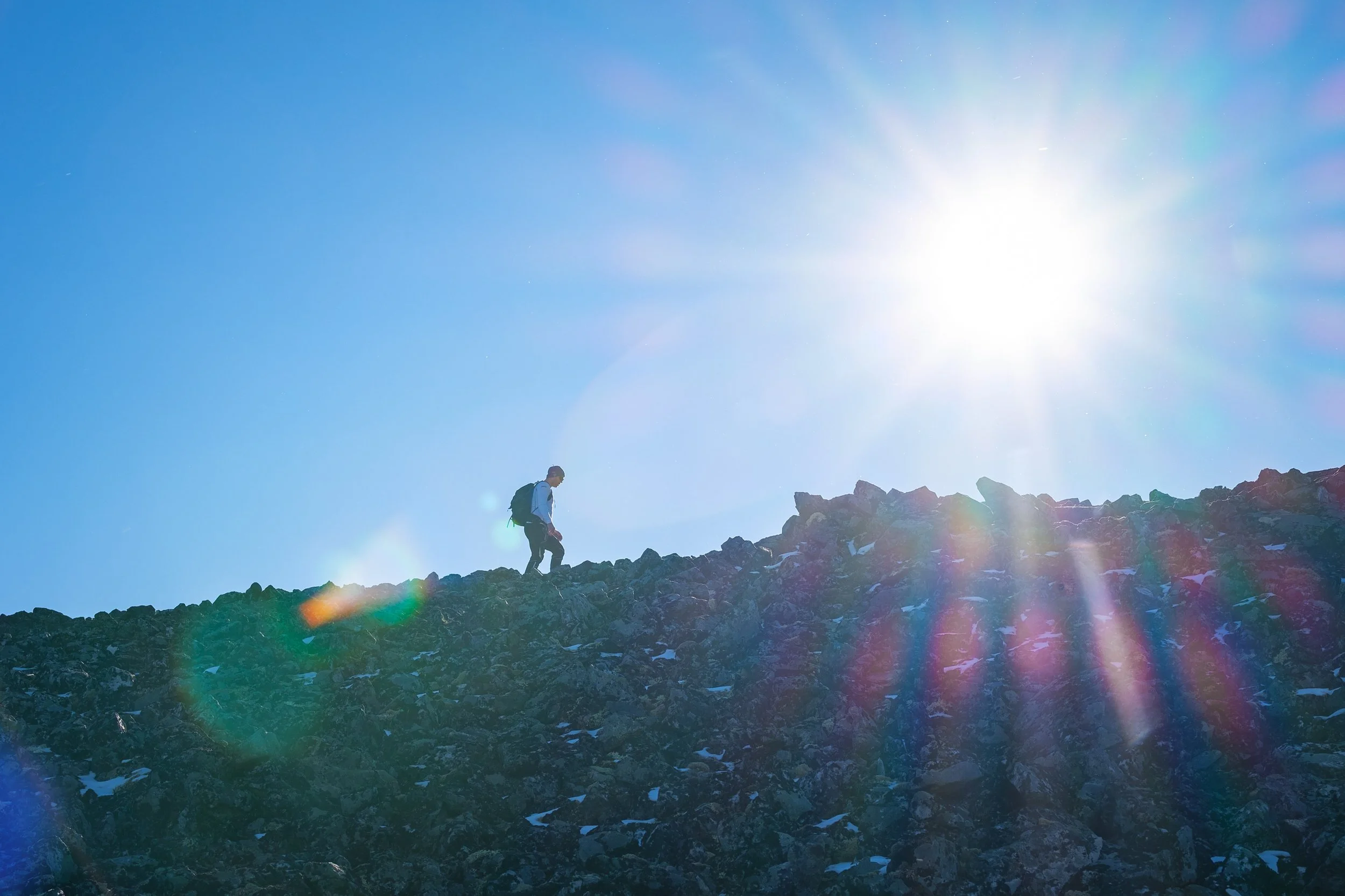 En person med ryggsekk klatrer opp en steinete fjellskråning under en klar blå himmel med sterk sol.