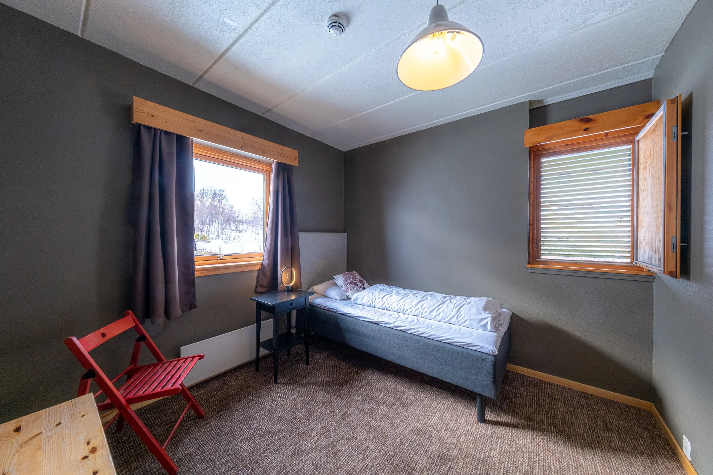 Enkeltrom med en enkelt seng, et lite bord, en rød sammenleggbar stol, to vinduer med trekarmer, beige gardiner, en taklampe, og nytt teppegulv.