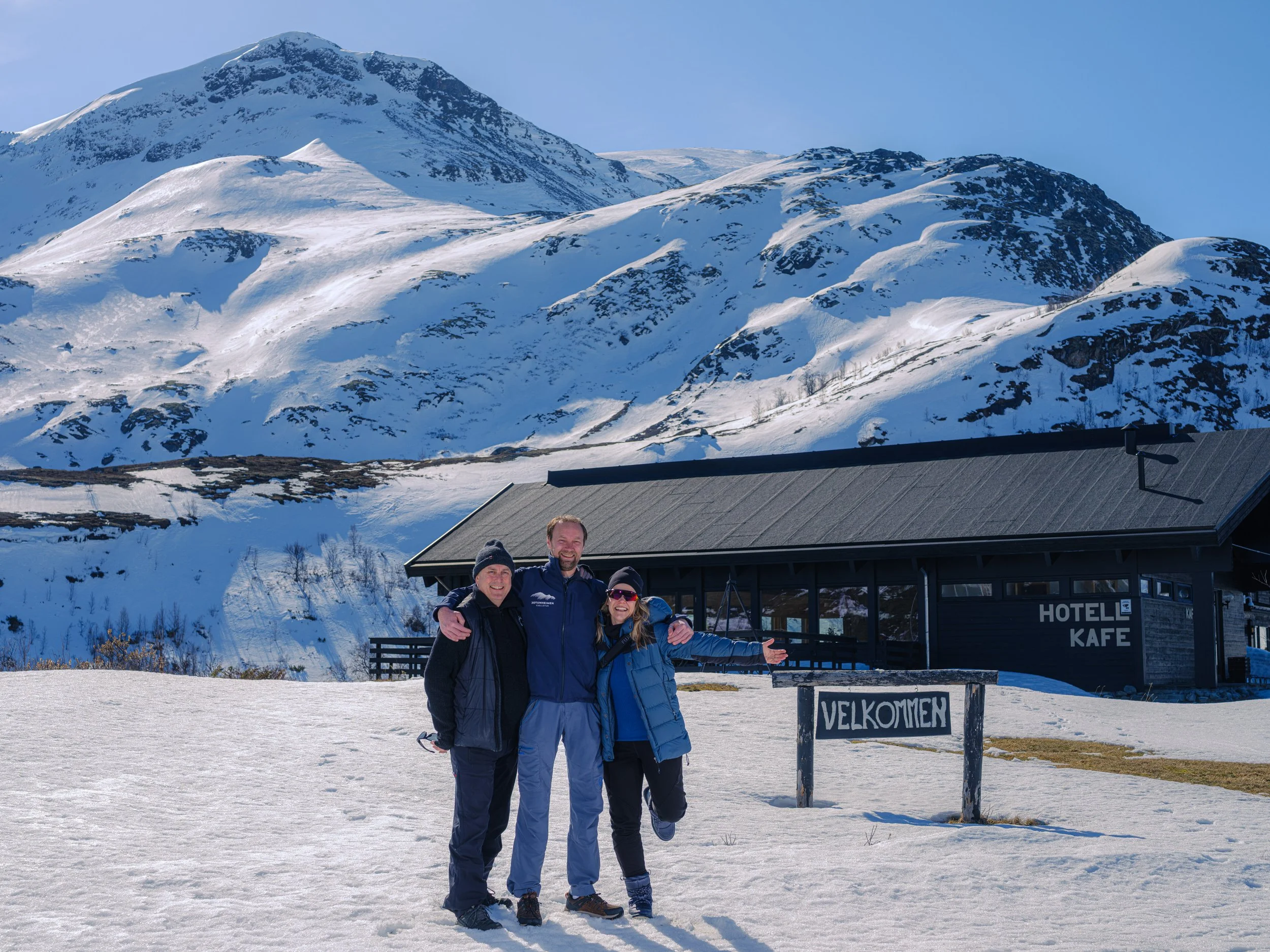 Tre personer står sammen i snø foran Jotunheimen Fjellstue med skilt 'HOTELL KAFE'. De smiler og har armene rundt hverandre. Bak dem er fjell med snø og klare blå himmel. En tavle med teksten 'VELKOMMEN' er i forgrunnen.