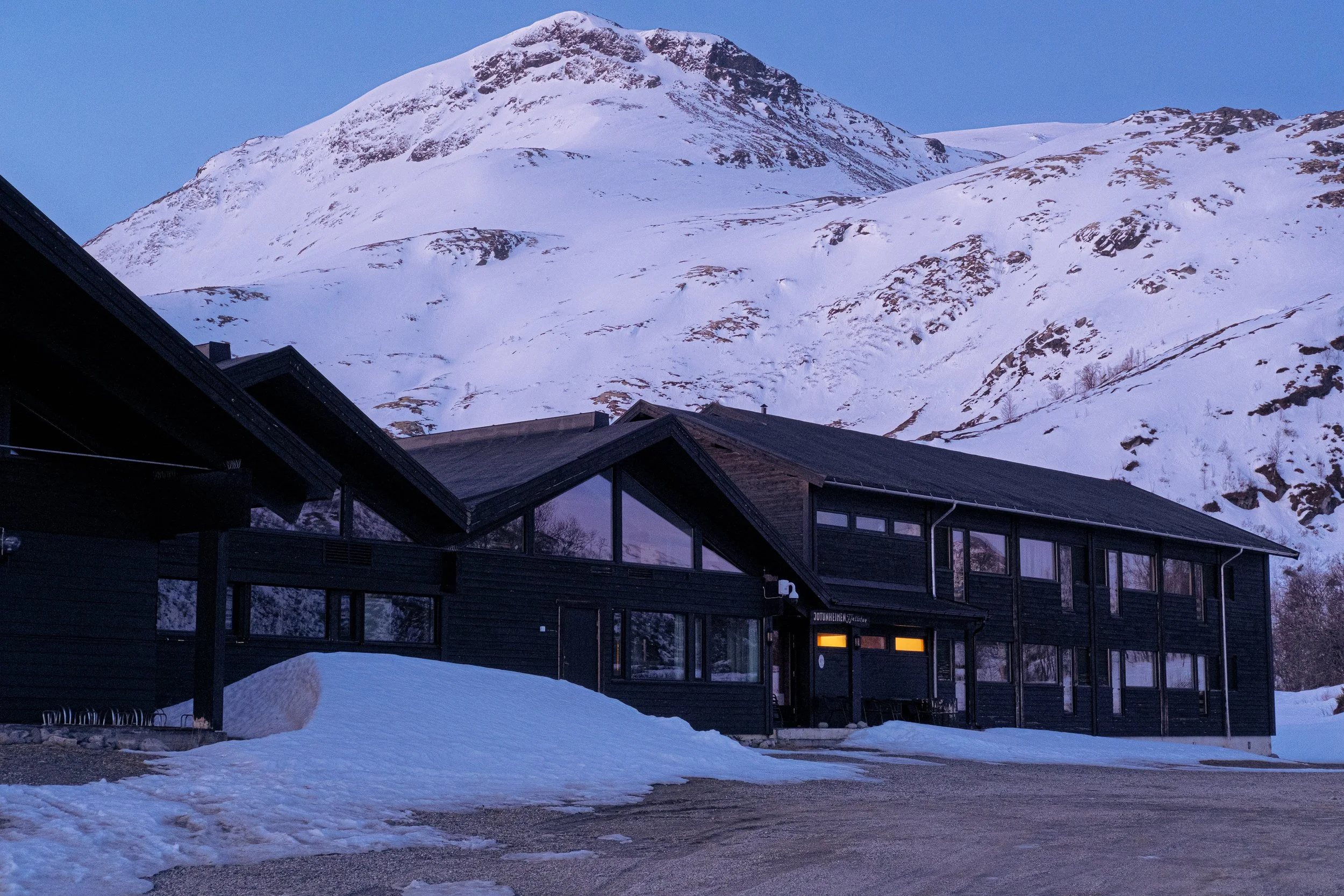 Jotunheimen Fjellstue med store vinduer omgitt av snø og plassert foran fjell med snødekke. Det er kveld og blåtime for solen går ned. 
