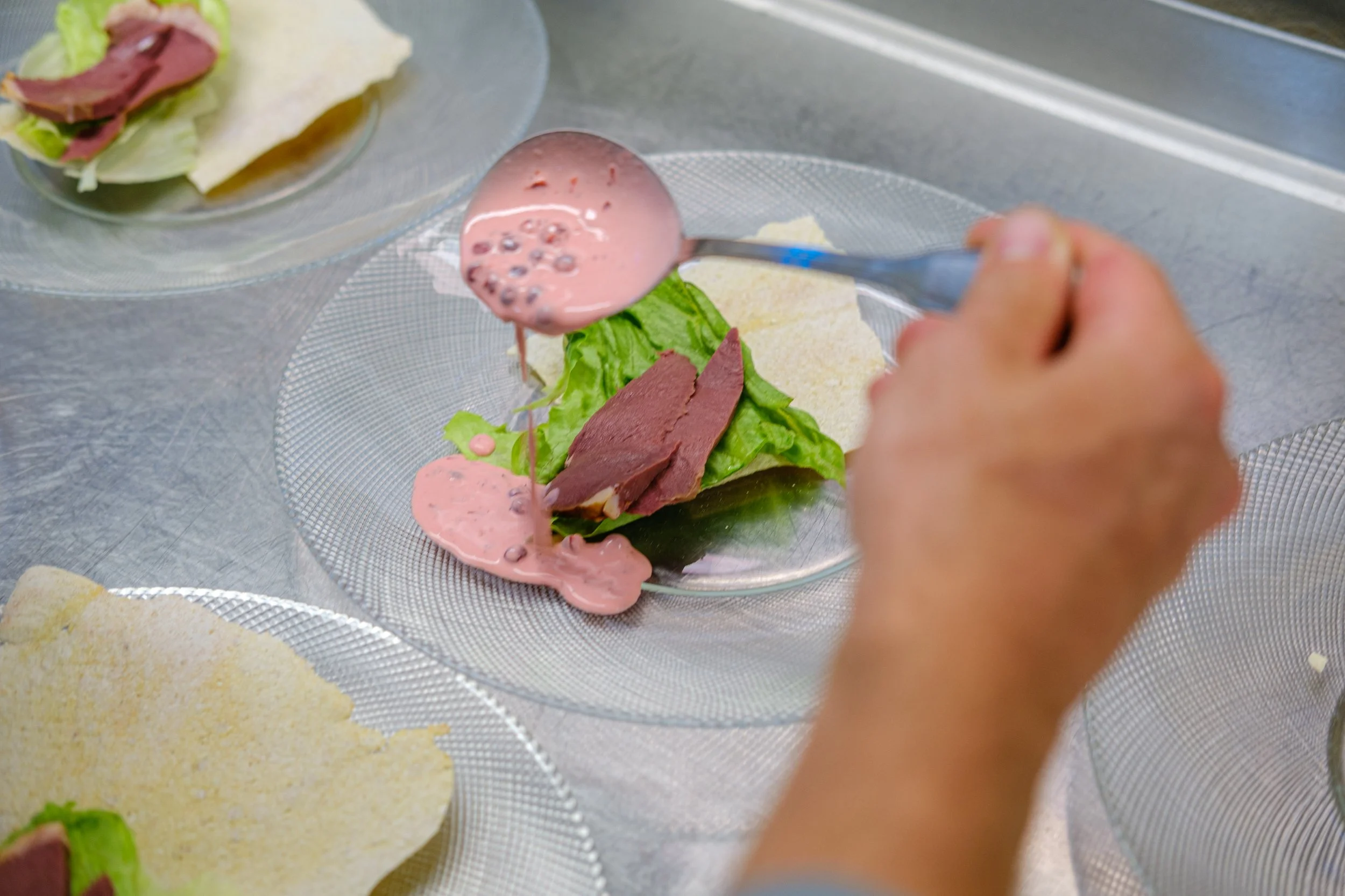 Kokk legger på saus på en tallerken med salatblader og skiver av kjøtt på en gjennomsiktig tallerken.