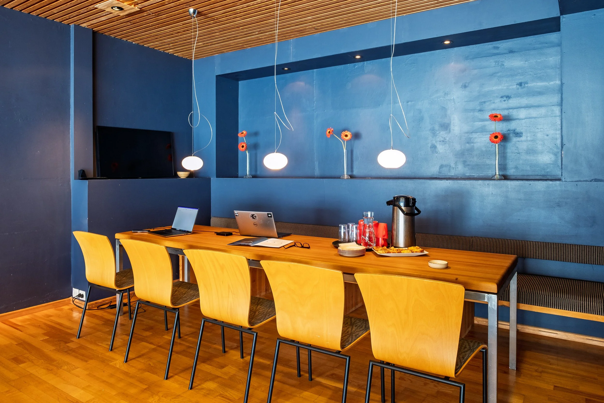 Møbler i møterom, inkludert en lang trebord, stoler, skjerm, en samling glass, en termos, tallerkener, og en servering av snacks, med blå vegger og tretak overtak. Dekorative vaser med solbrune blomster henger fra taket.