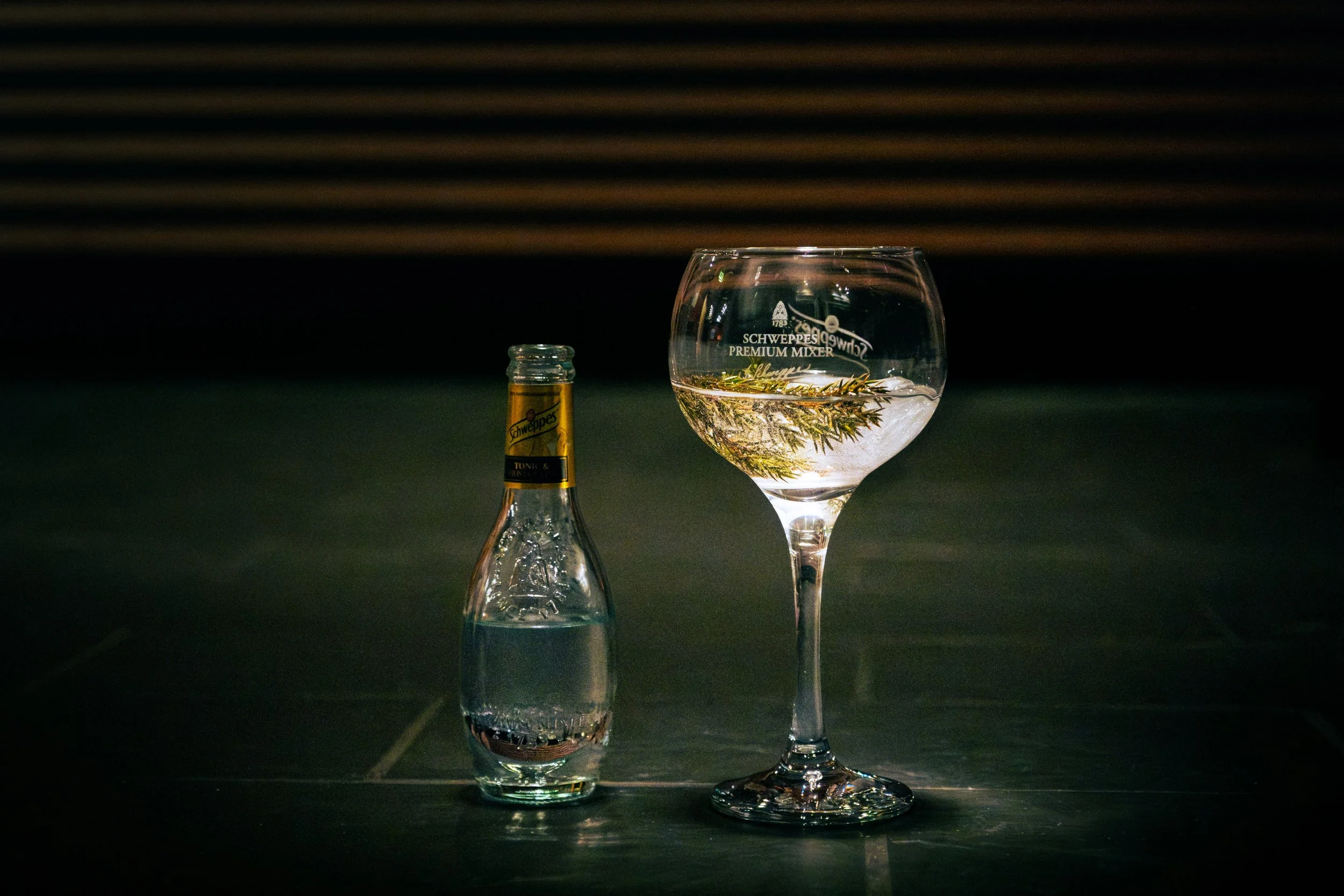 En åpen liten flaske med klar væske ved siden av en større glass med stilk, fylt med klar væske og en kvist med grønne blader.