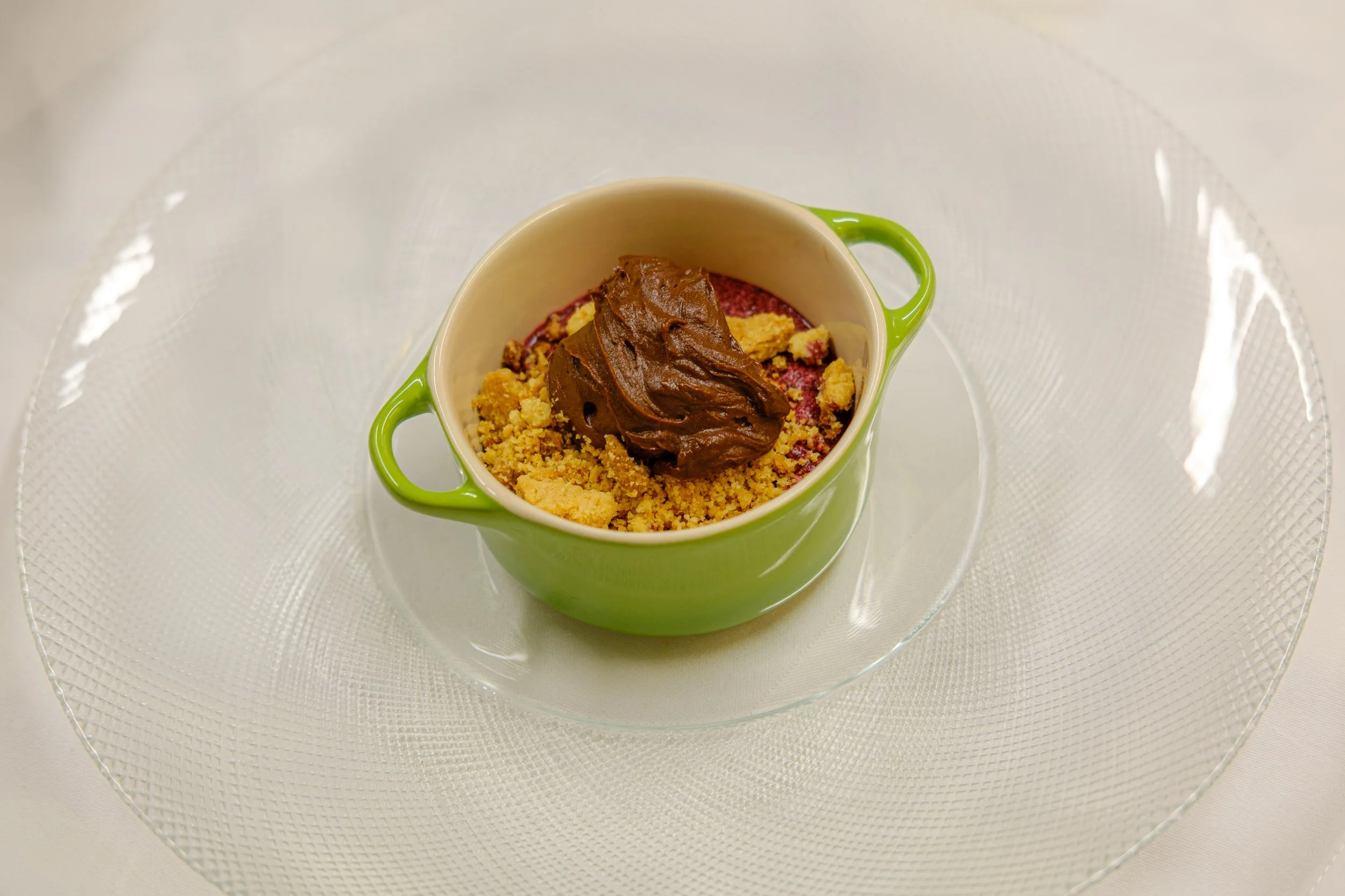 Dessert i en grønn kopp med sjokolade og sprø topping, plassert på en glassplate.