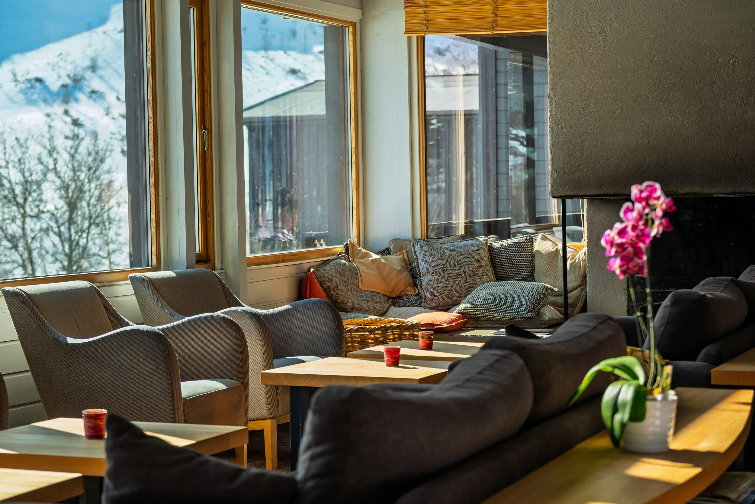 Innendørs stue med store vinduer som viser snødekt landskap ute. Møbler inkluderer lenestoler, et hjørne med mange puter, og et bord med lys.