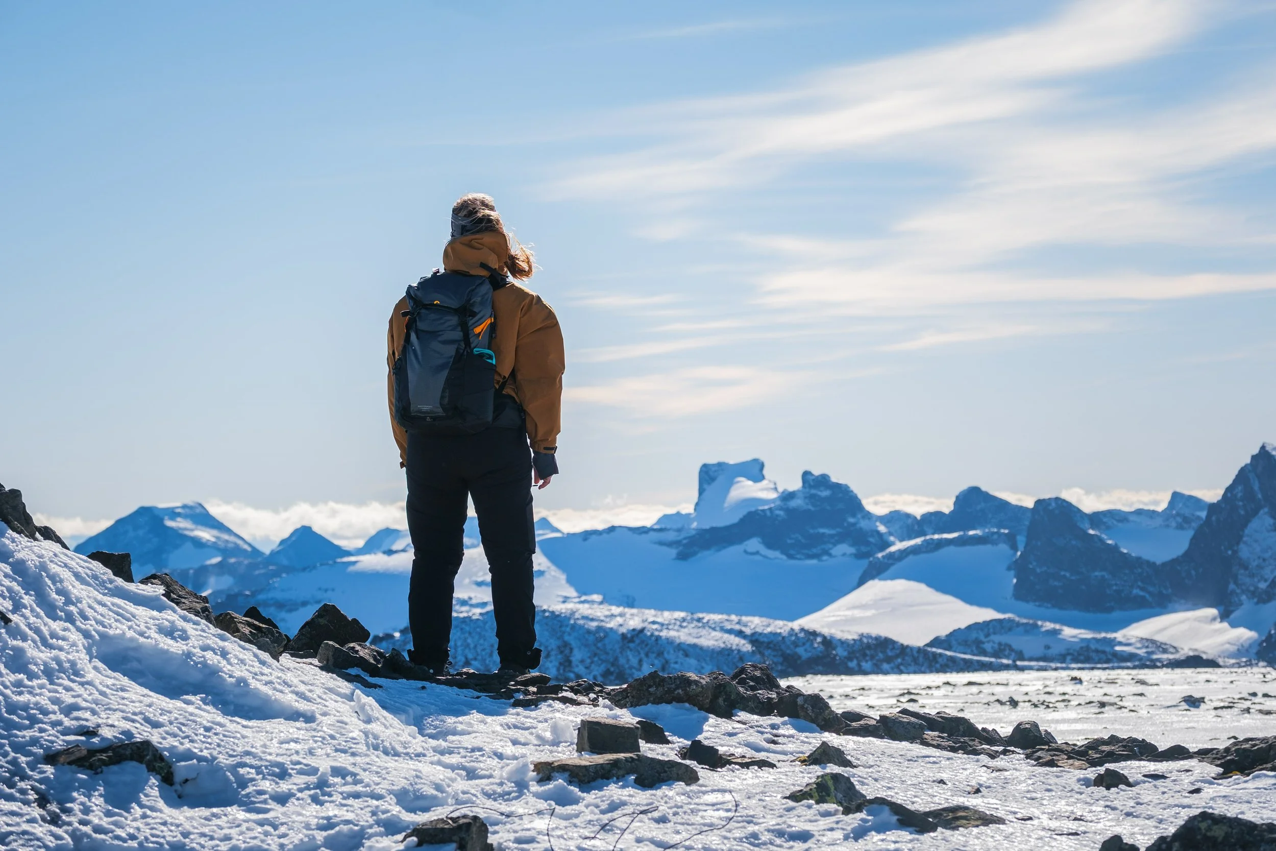 En person med ryggsekk står på snødekt fjell under klar himmel med snødekte fjell i bakgrunnen.