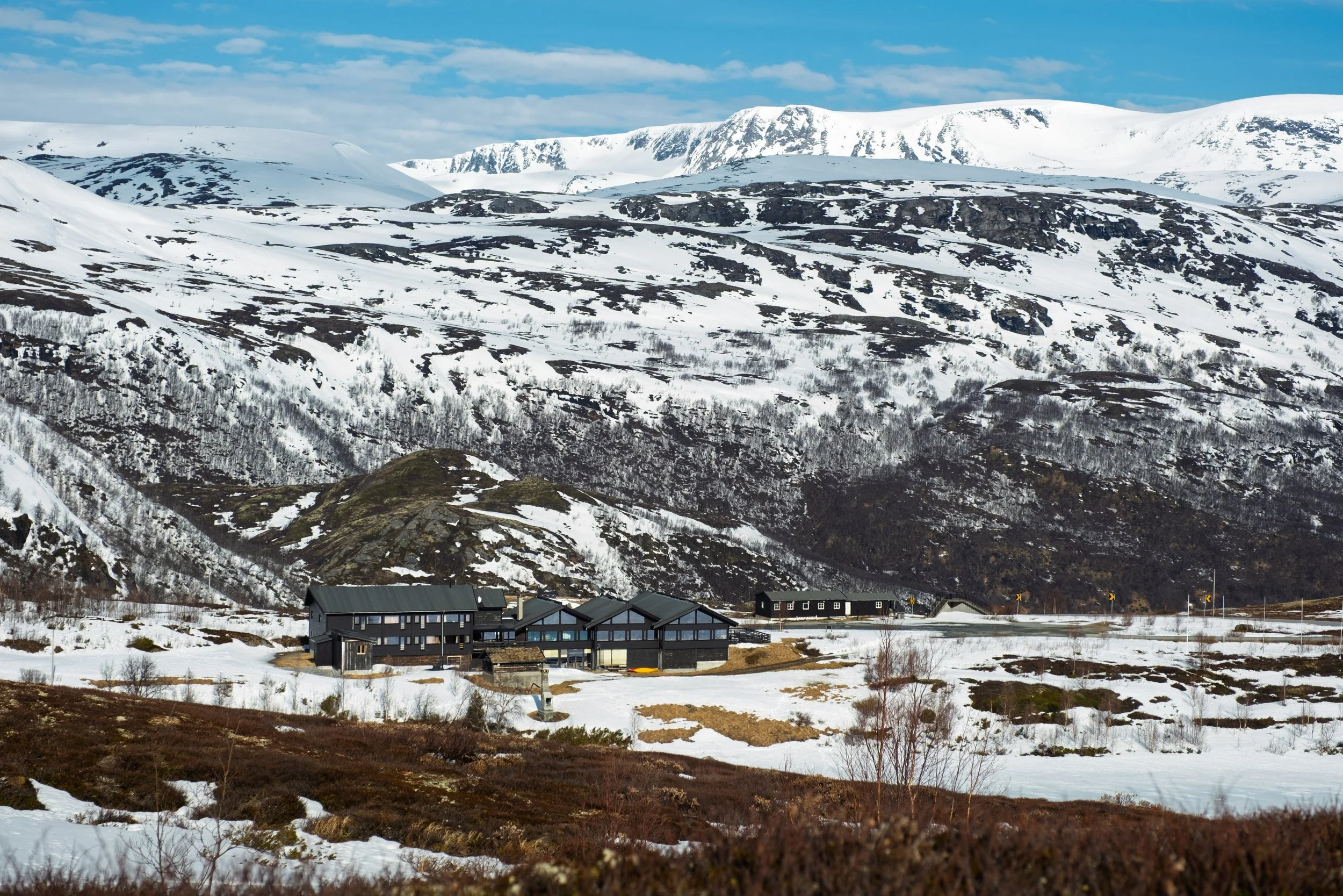 Jotunheimen Fjellstue med svarte bygninger i snødekt fjellandskap under en blå himmel.