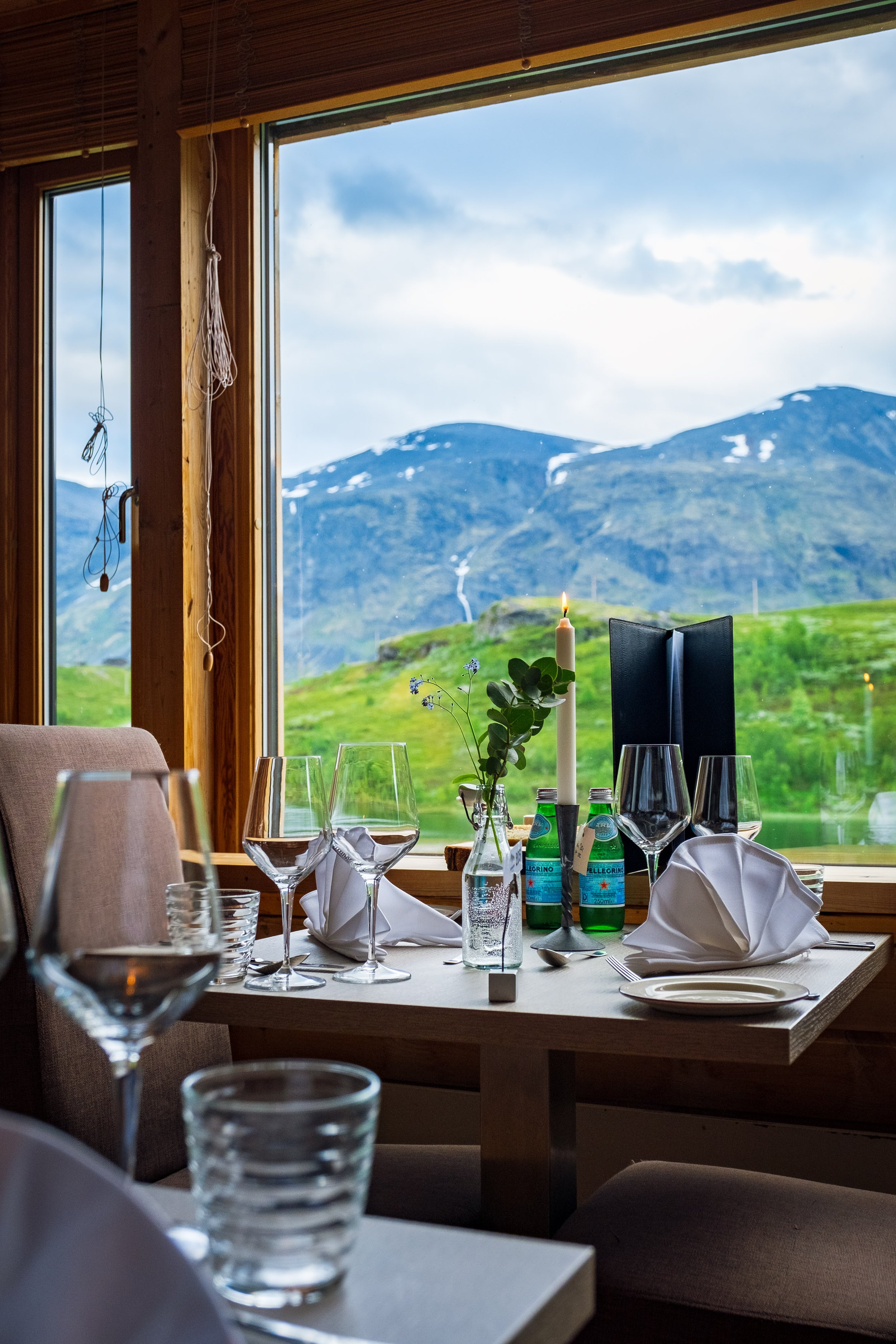 Inne i en restaurant med utsikt over grønne fjell og snødekte fjelltopper, satt opp til middag med tomme tallerkener, glass, bestikk, lys, og blomster.
