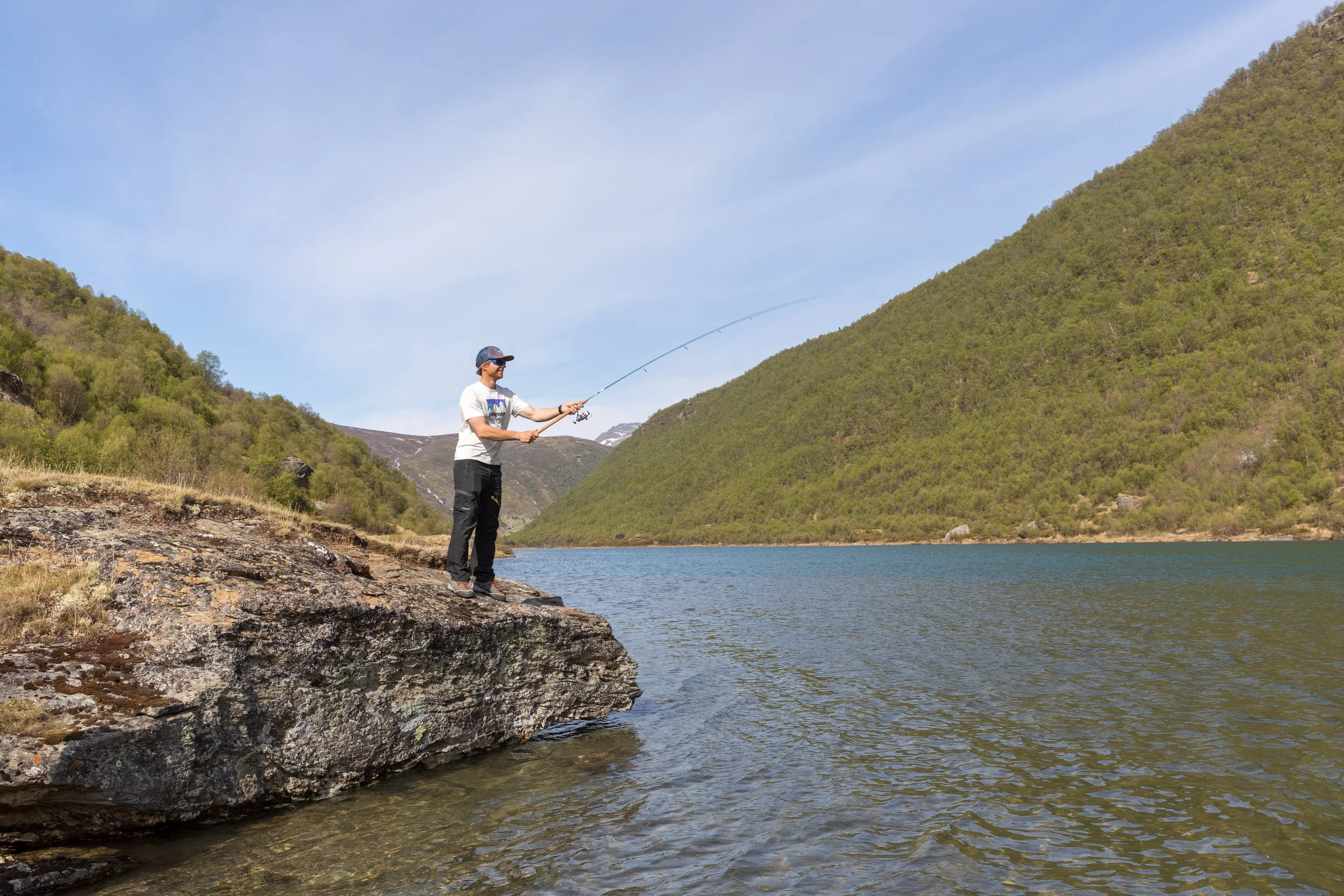 En mann står på en stein ved vannet og fisker med en fiskestang i et Jotunheimen med grønne fjell og blå himmel.