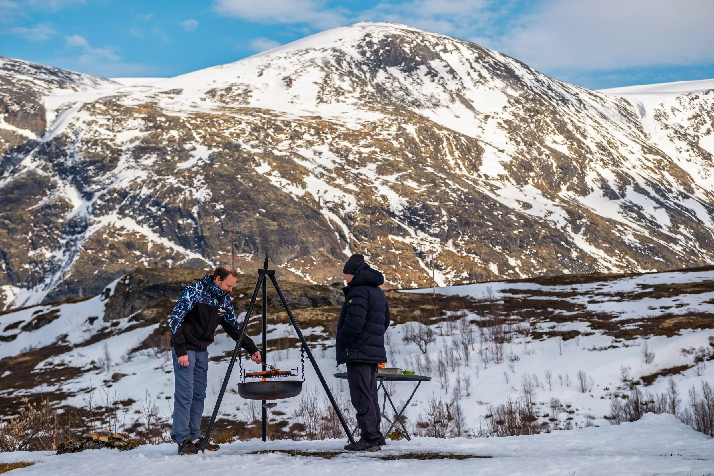 To personer står ved en bålpanne i snødekt fjellandskap, med høye fjell og en blå himmel i bakgrunnen.