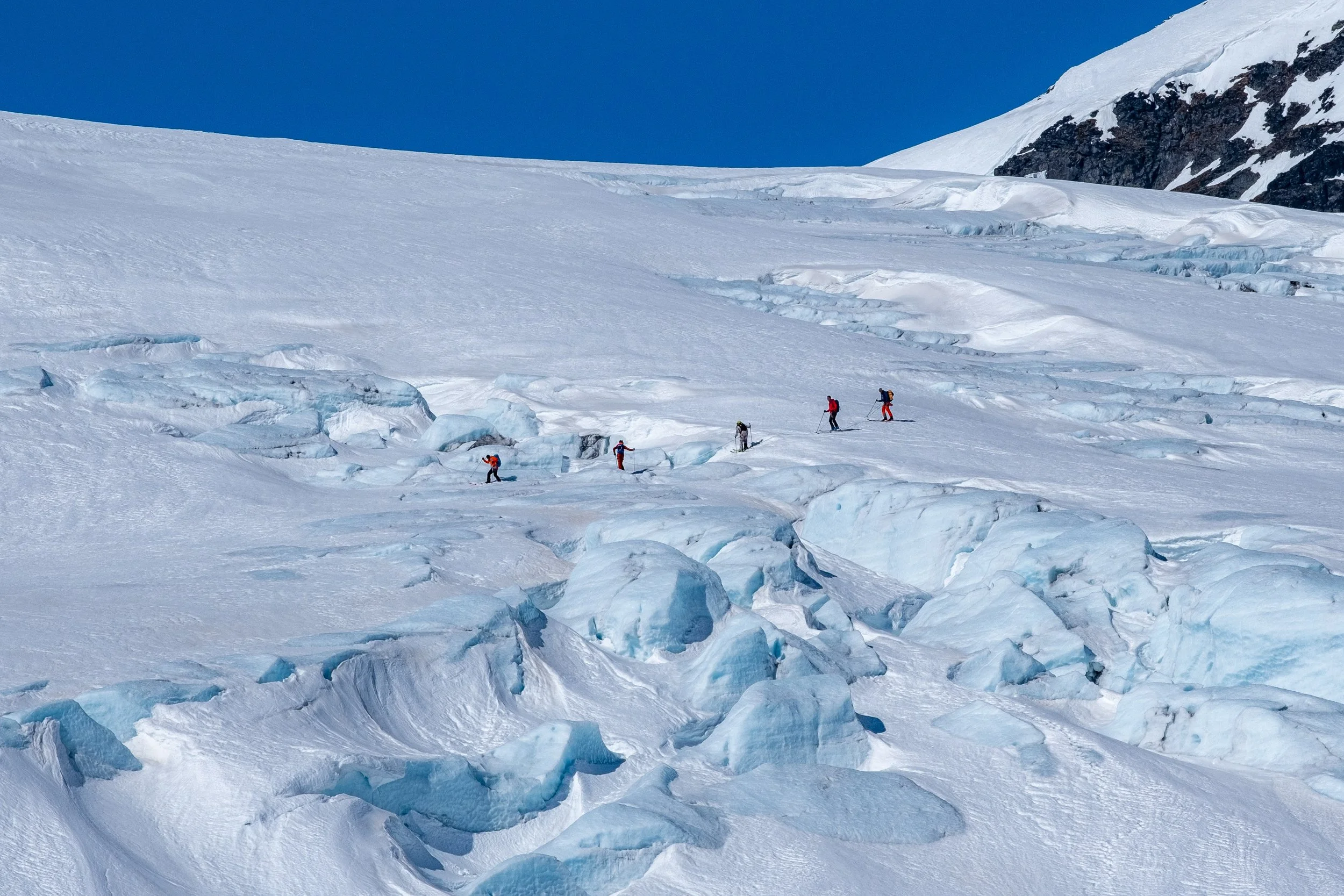 Fjell med snø og isbre med seks personer som går på ski.