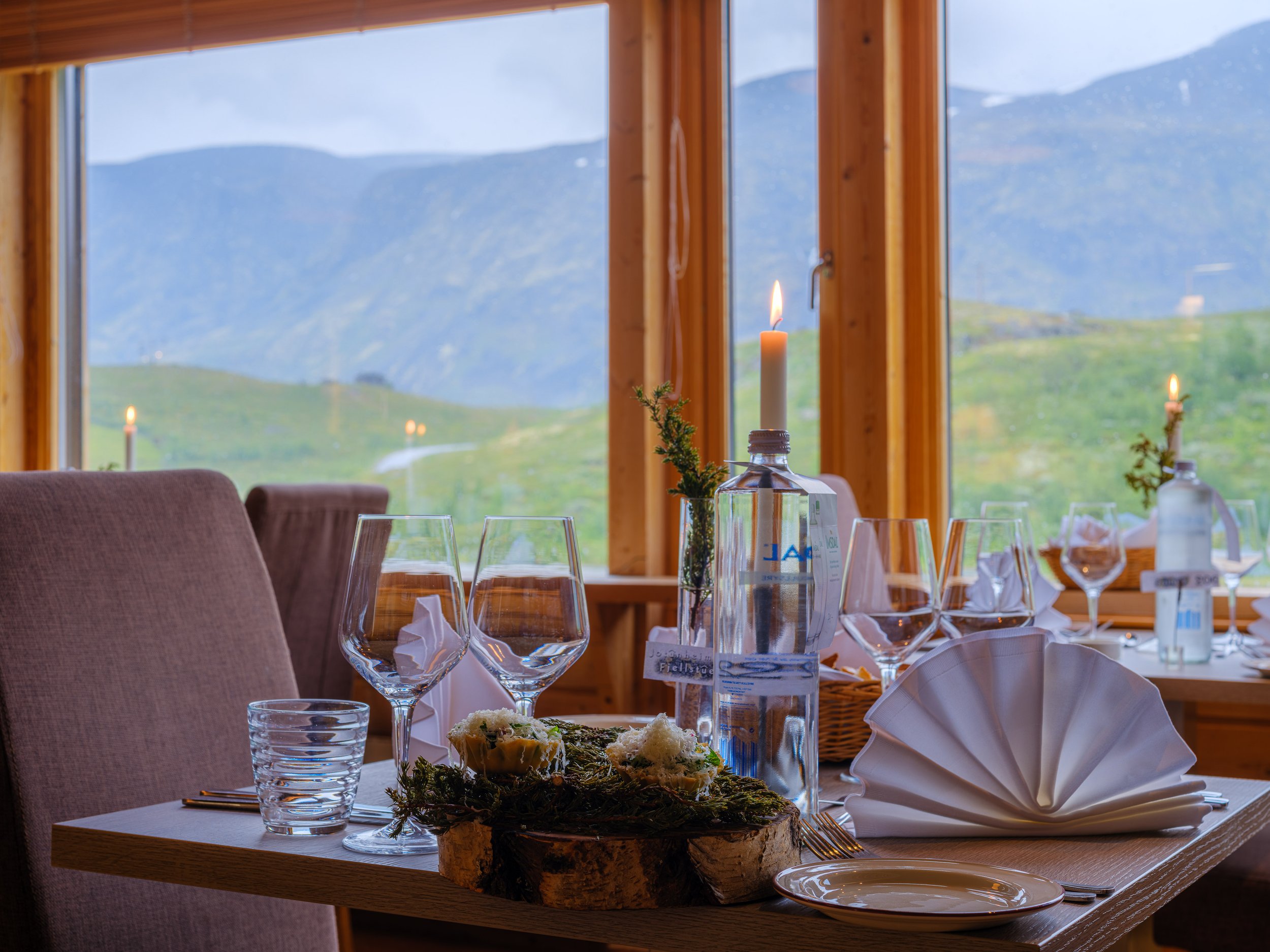 Elegant middagsbord dekket med glass, tallerkener, servietter og stearinlys i rustikk restaurant med fjellutsikt.