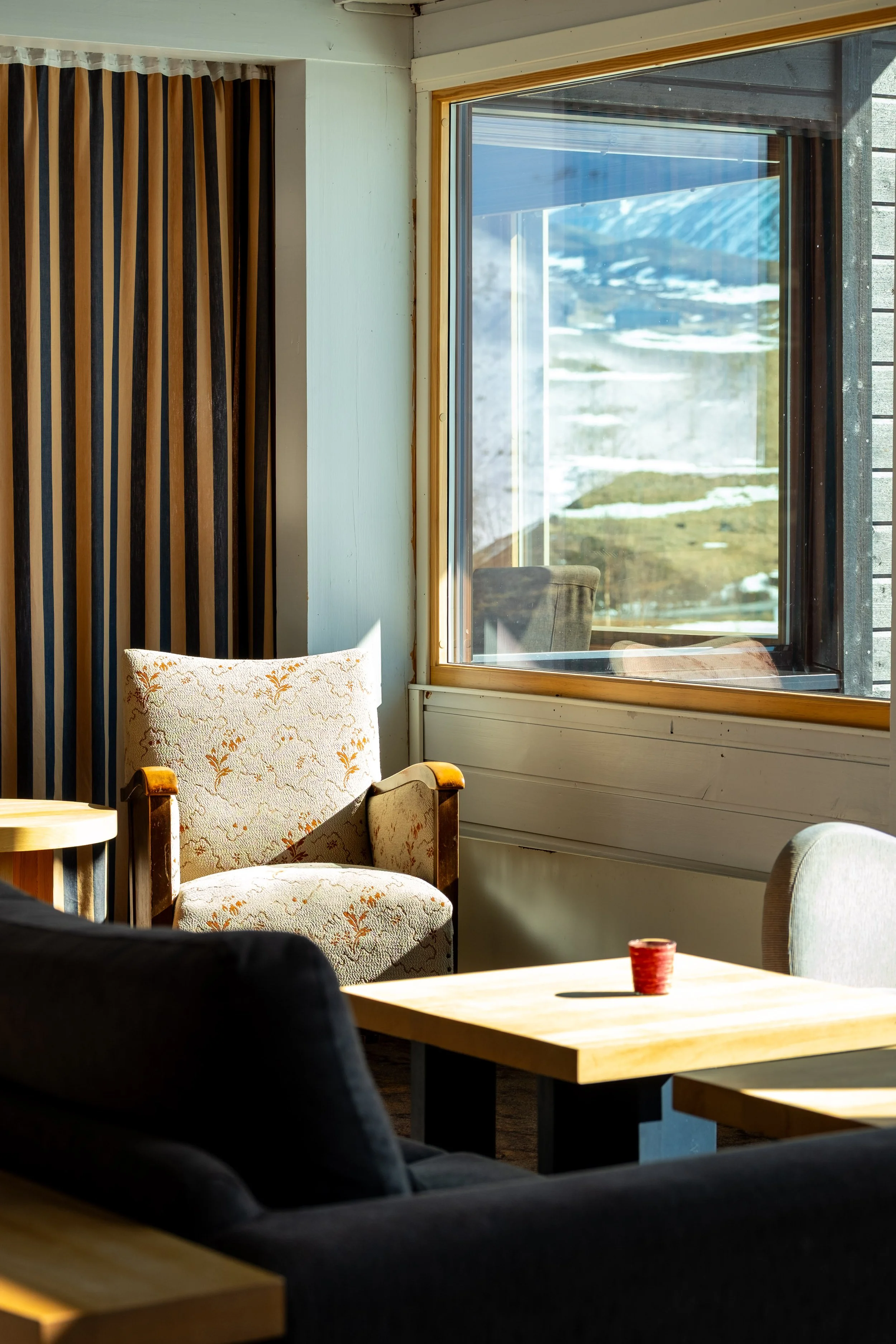 Innendørsområde med et trestolssete, en sofa, et trebord med tente lys og en vindusrute som viser en naturlandskap utenfor.