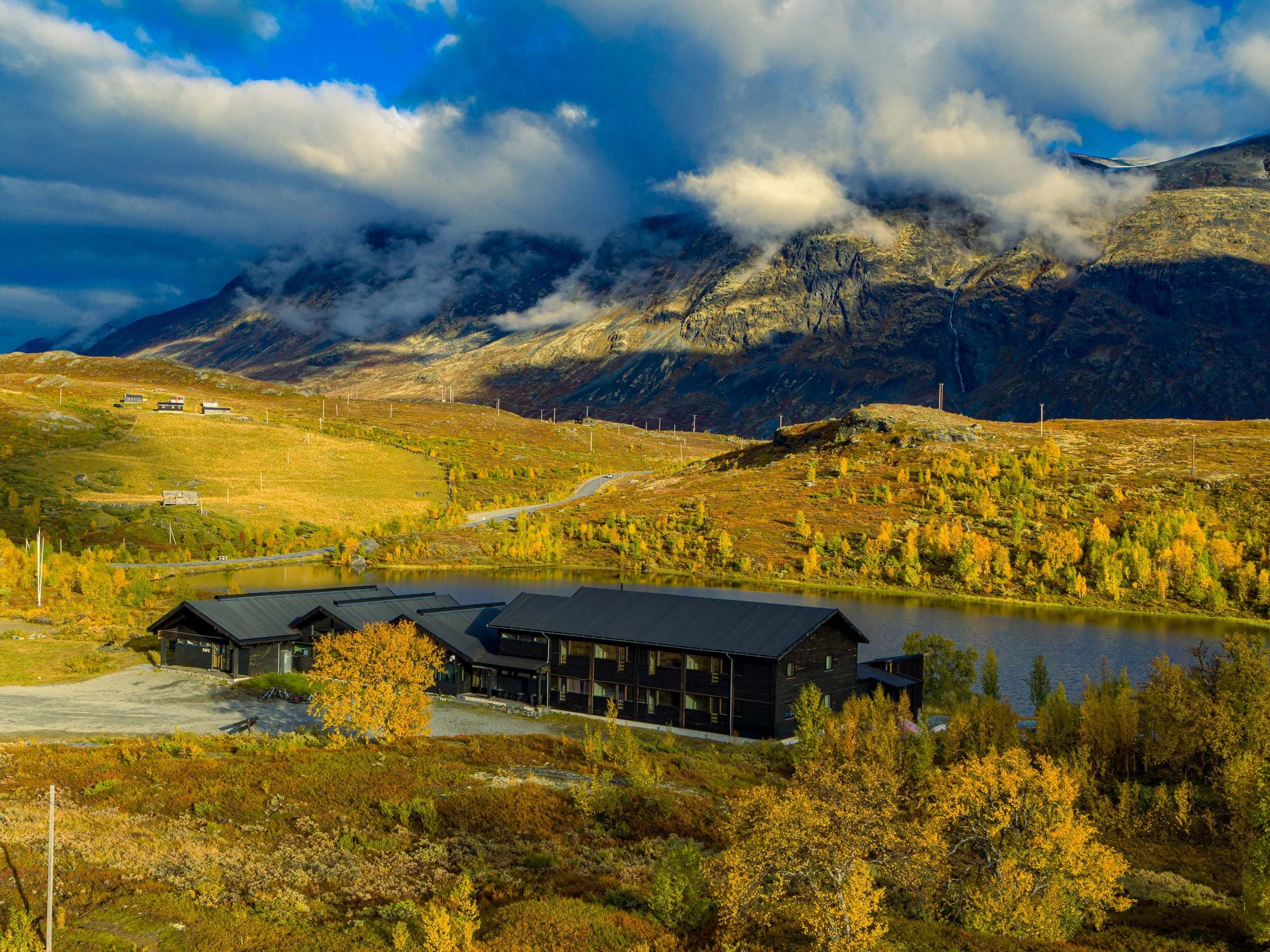 Høstlig landskap med fjell, en innsjø, og hotellet, Jotunheimen Fjellstue, i forgrunnen, omgitt av fargerike trær.