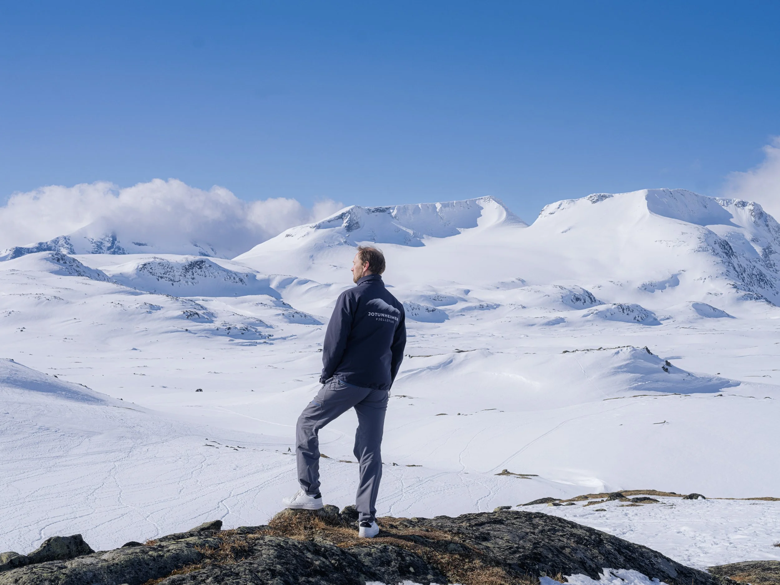 Mann står på en stein oppå snødekte fjell med fjellkjede i bakgrunnen, blå himmel og noen skyer.