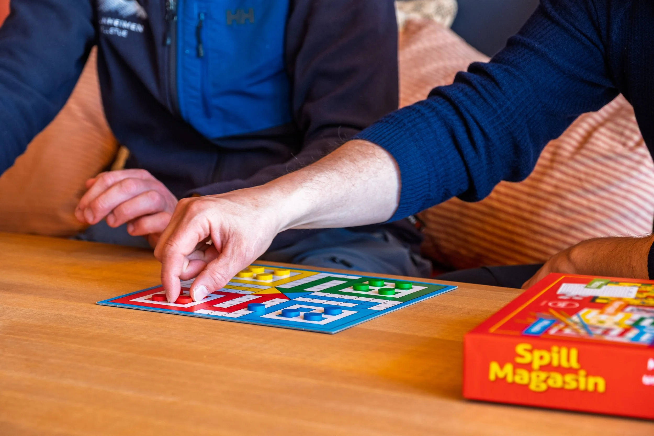 To personer spiller Ludo ved et bord. En person er i ferd med å plassere en rød brikke, mens det ligger andre brikker i ulike farger på brettet. 