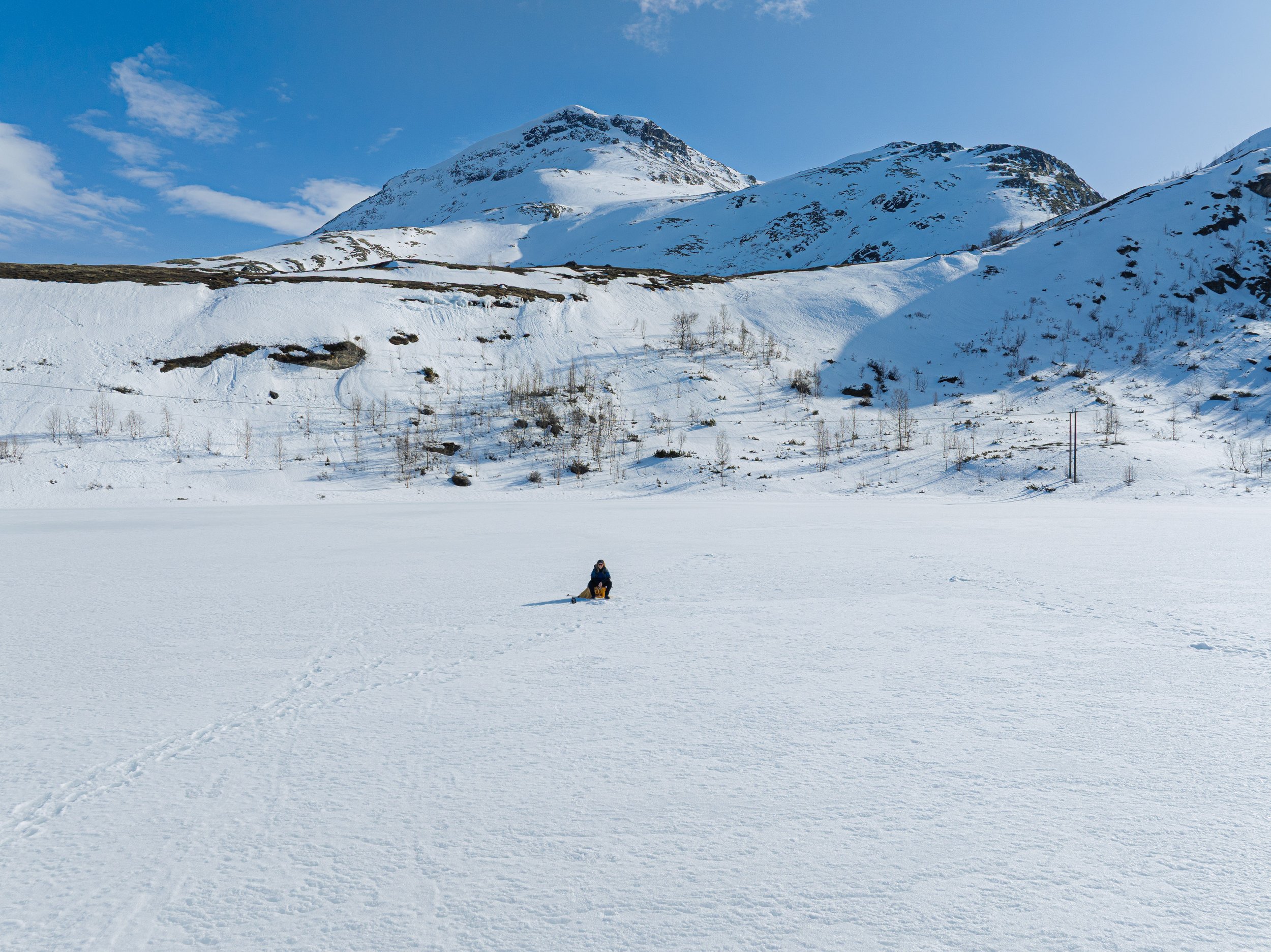 En person som sitter og isfisker med fjell i bakgrunnen under en klar blå himmel.