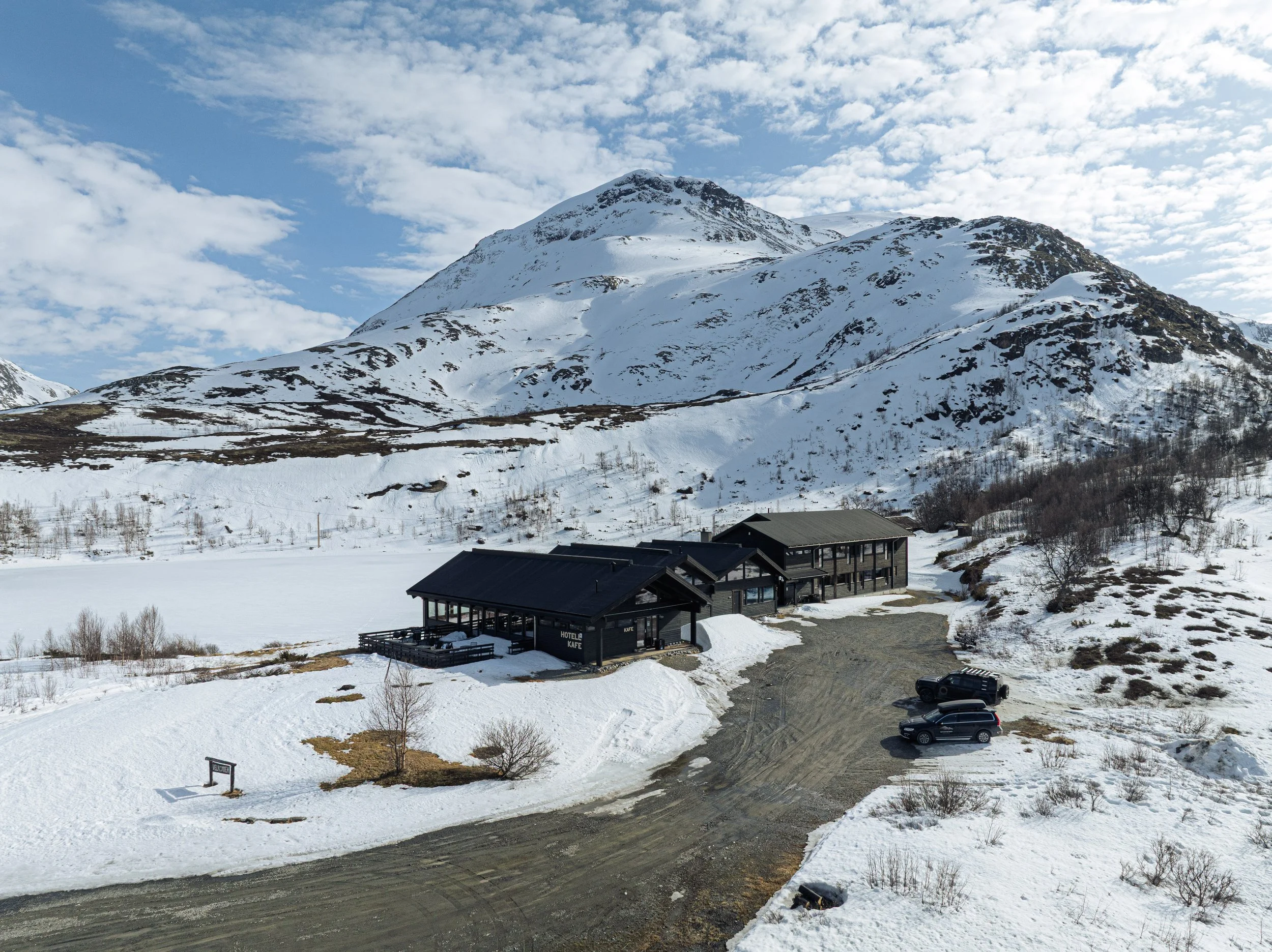 Snødekt fjellandskap med Jotunheimen Fjellstues bygninger og parkering i forgrunnen, omgitt av snødekte trær og fjell i bakgrunnen under en skyet himmel.
