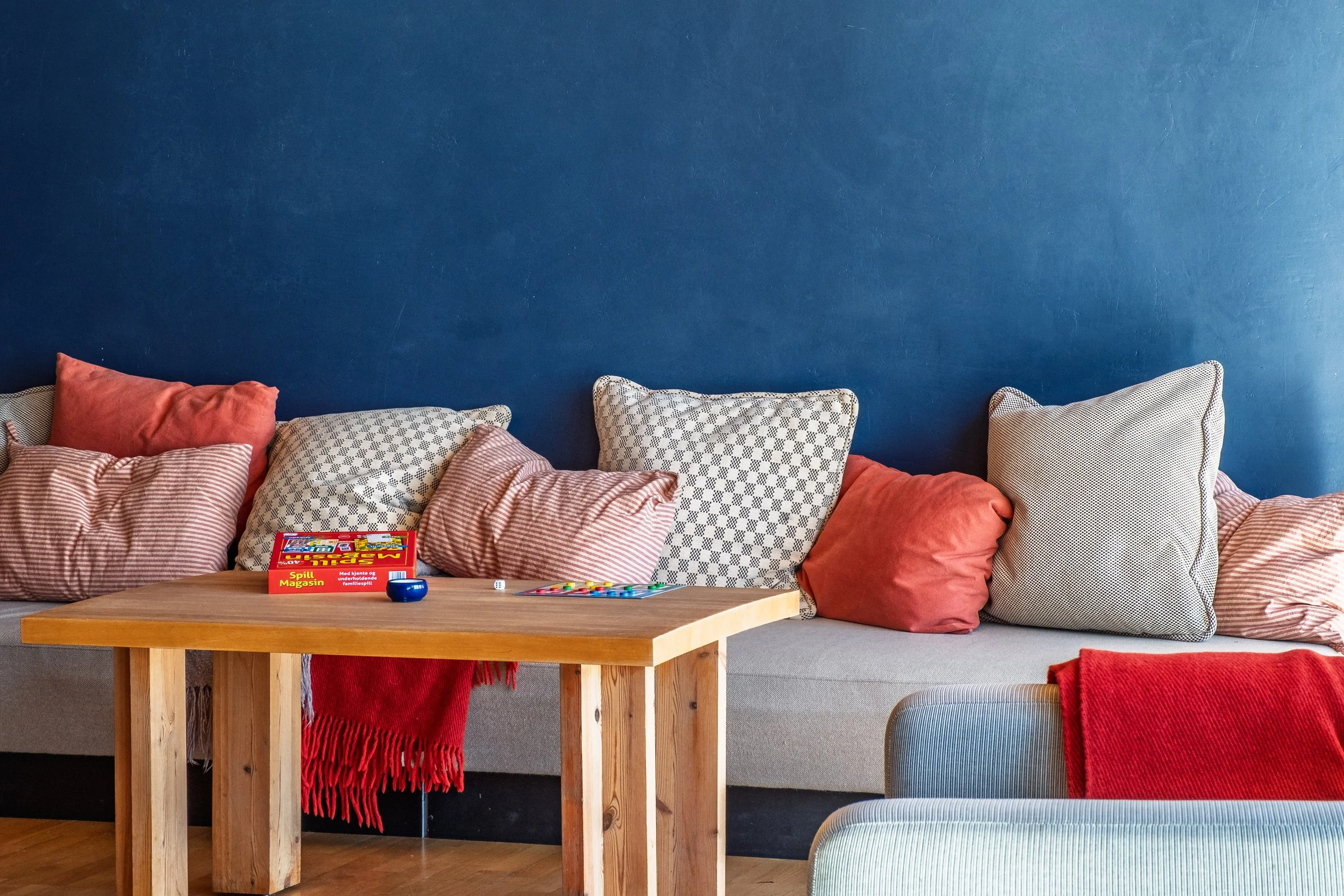 Stue med en lang sofa med mange puter i ulike mønstre og farger. En enkel trebord med et spill og små gjenstander på. 
