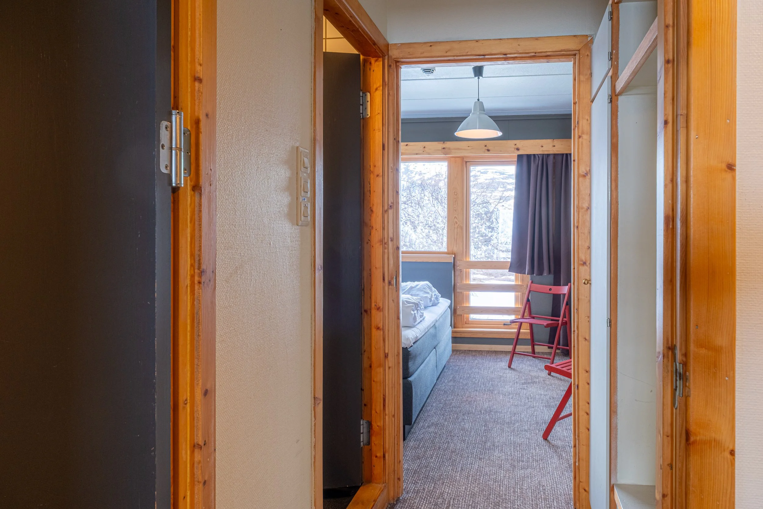 Utsikt inn til soverom med stor vindu, dobbeltseng, rød stol, mørke gardiner og treverk på vegger og rammer.