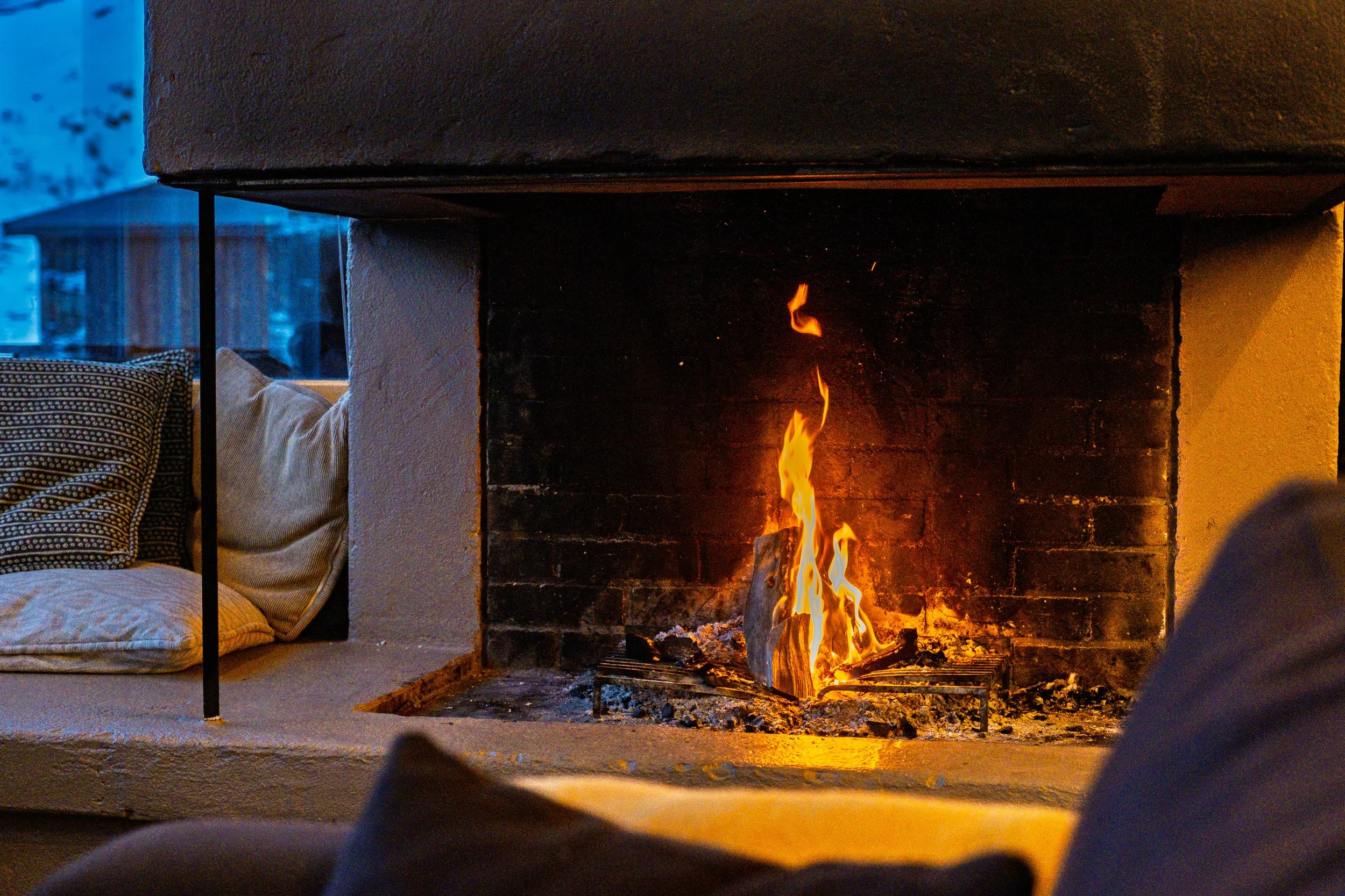 En peis med brennende ild, omgitt av en hvit murvegg, med puter i et sittehjørne. 