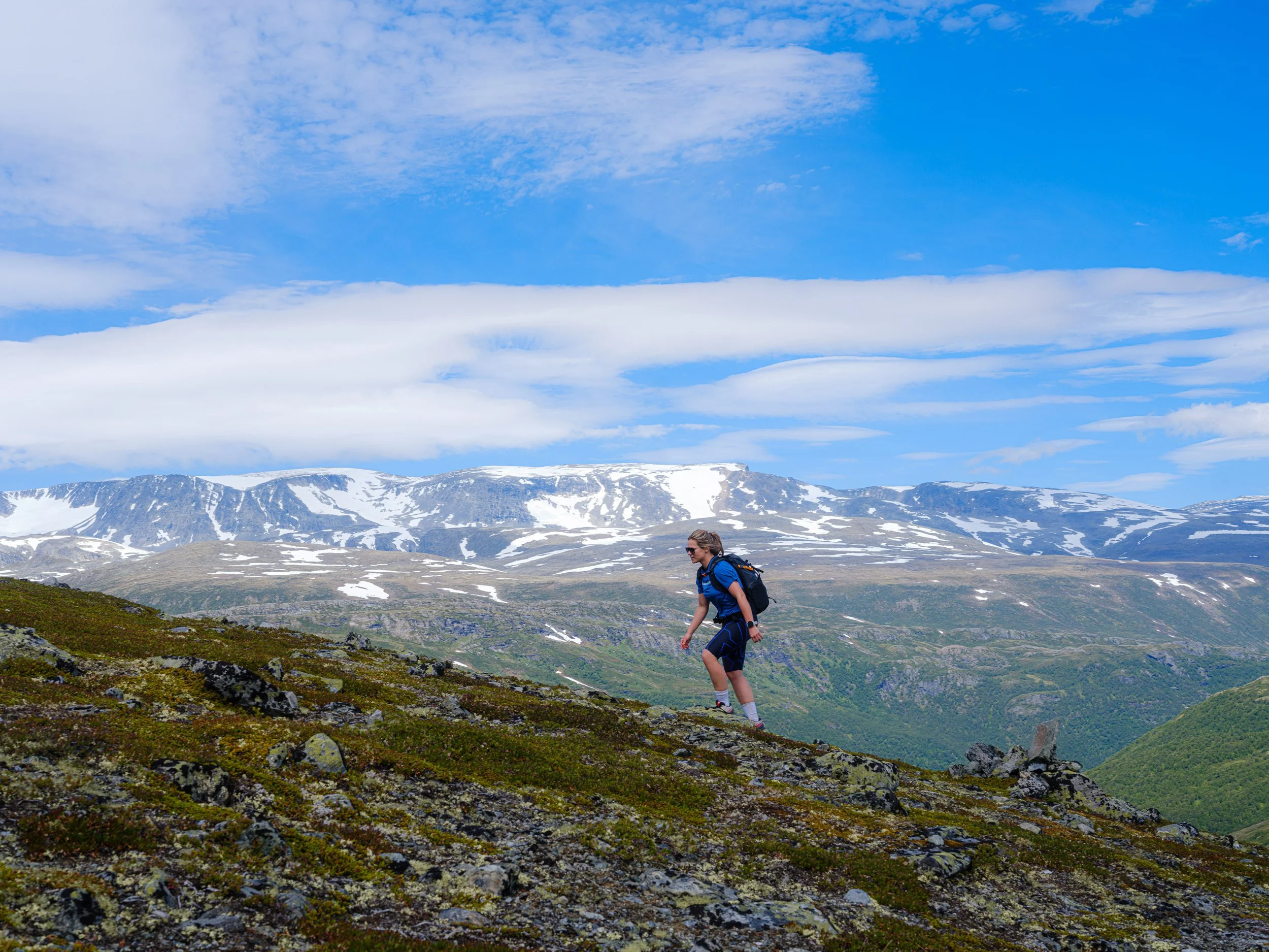 En person går oppover i fjellandskap med snødekte fjell og blå himmel med spredte skyer i bakgrunnen.