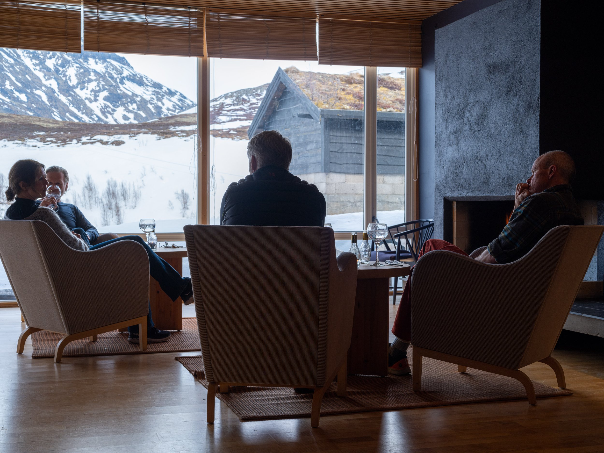 Fire personer sitter i en peisestue med store vinduer som viser snødekte fjell og naturlandskap. De drikker drikke og snakker.