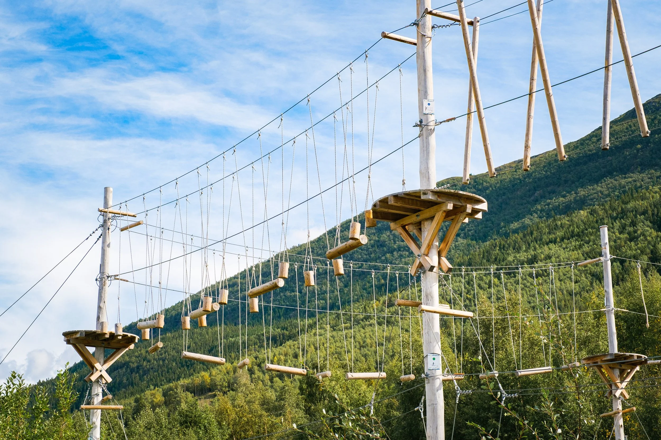 Utendørs klatreopplegg med tau og treplattform mot en bakgrunn av grønne fjell og blå himmel.