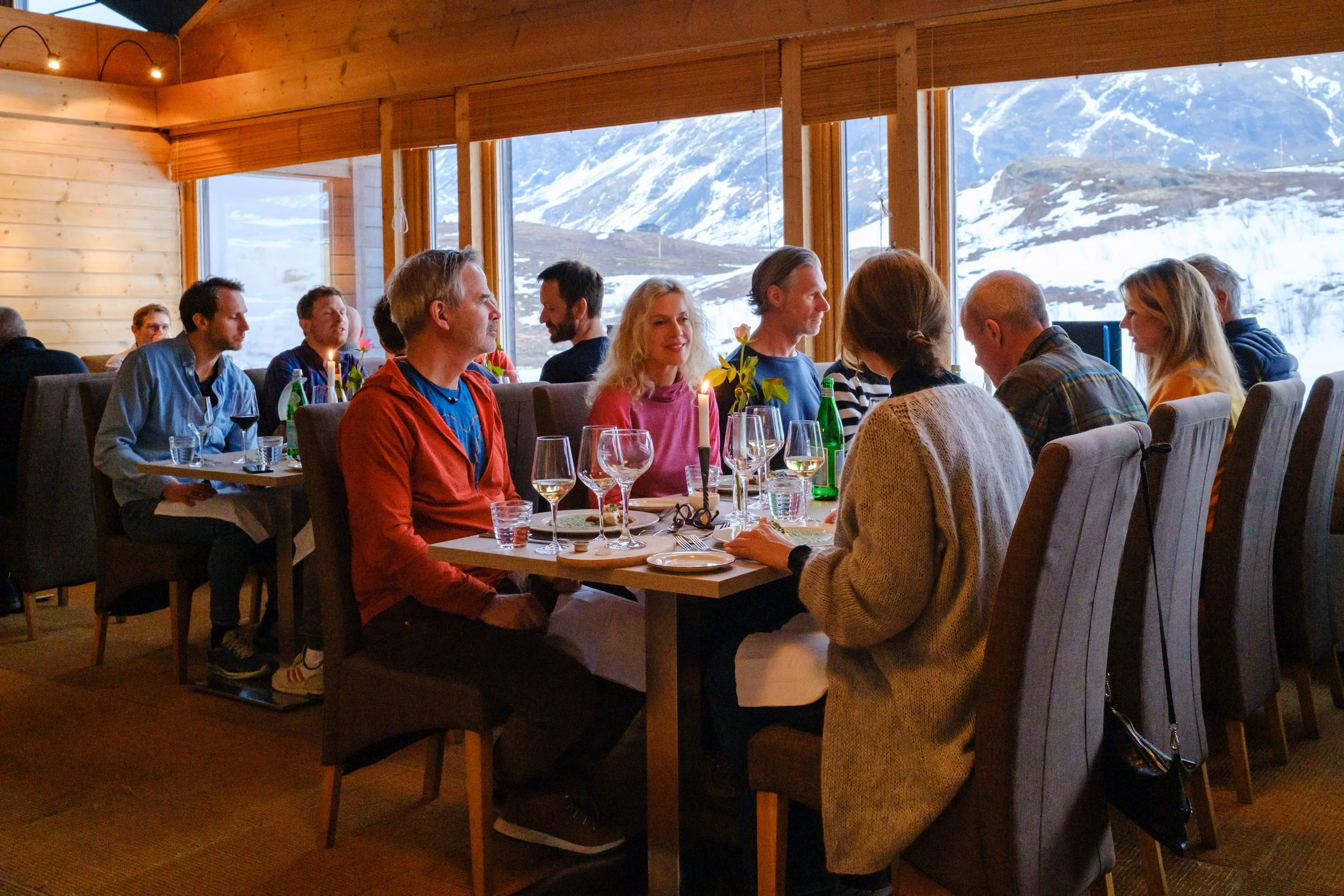 Gjestemiddag med personer som spiser og snakker i et restauranten med store vinduer som viser snødekte fjell.