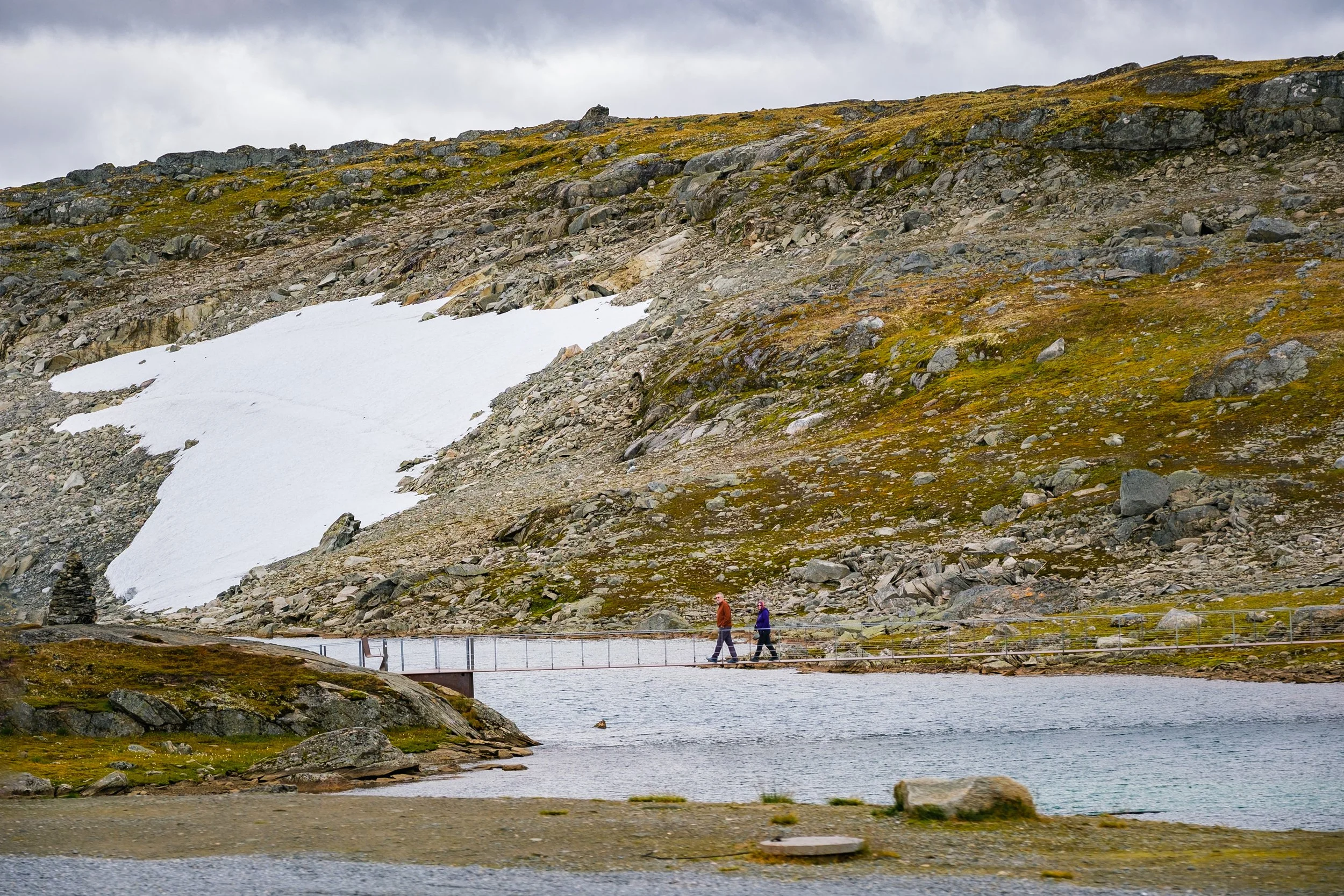 To personer går over en bro over vann omgitt av fjell med snø og fjellvegetasjon.