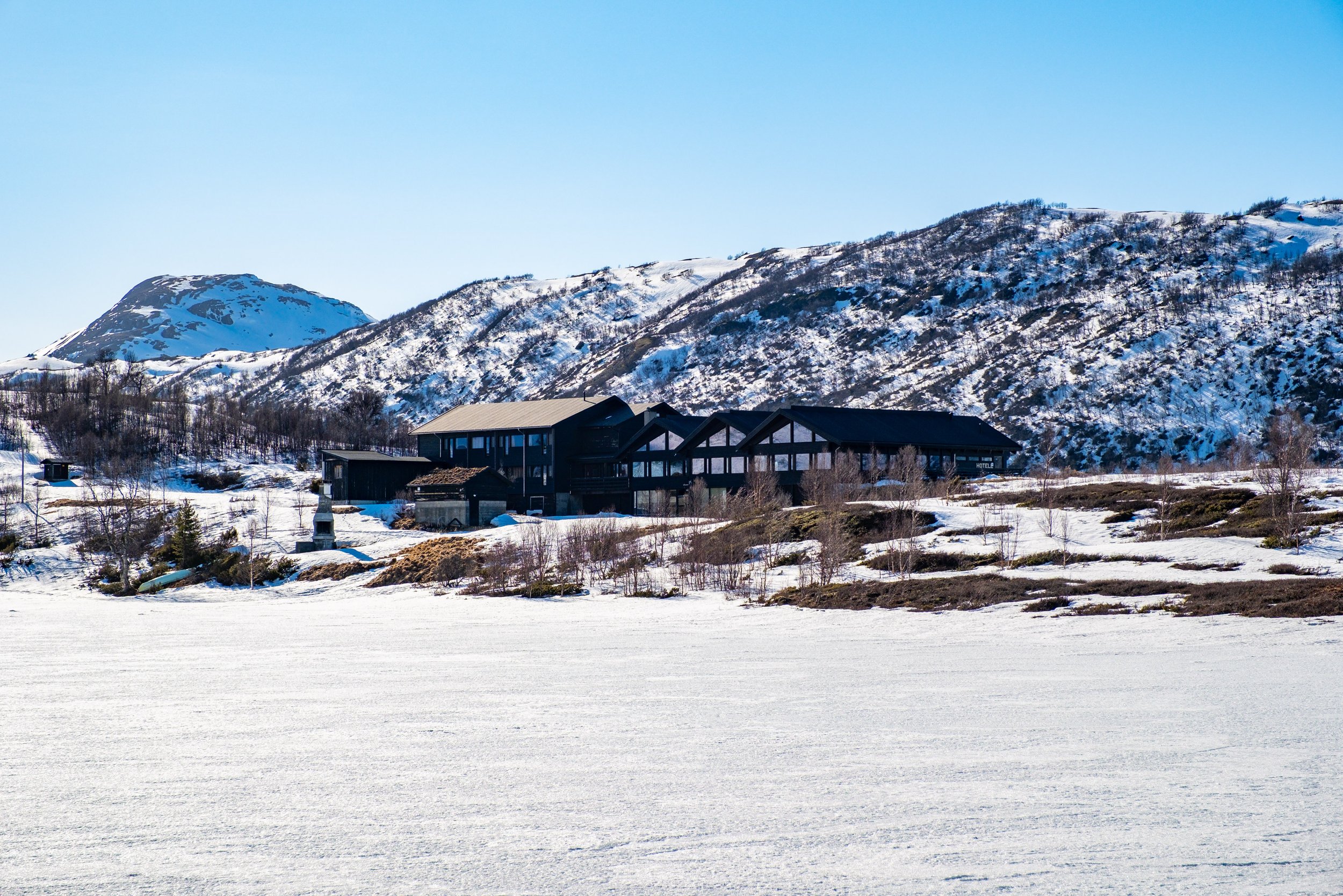 Snødekt fjellandskap med Jotunheimen Fjellstue i midten, omringet av små trær og snøfylte områder under en klar blå himmel.