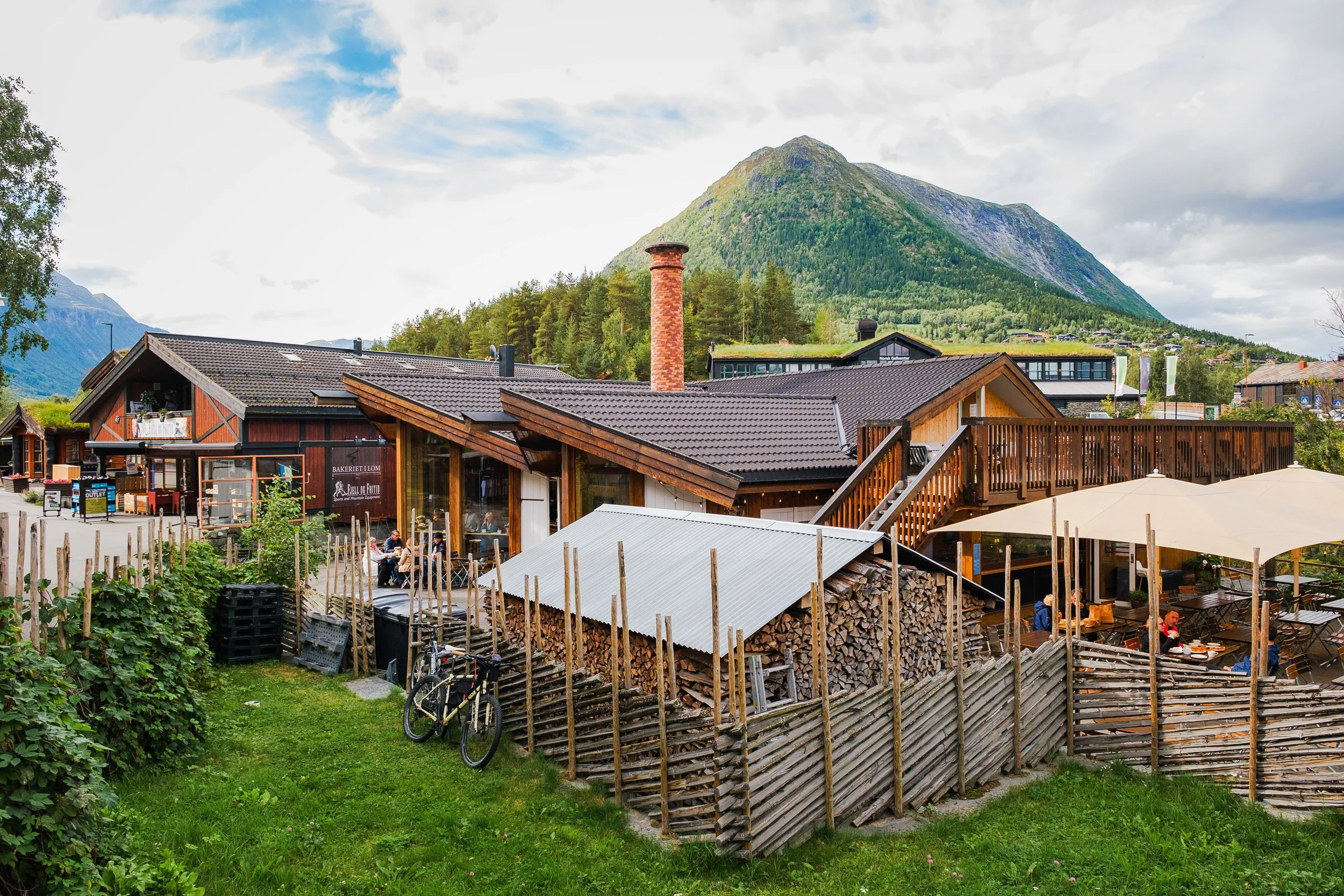 En liten restaurant og Lom bakeri fjellområder, omgitt av trær og med fjell i bakgrunnen, med utendørs sitteplasser. 