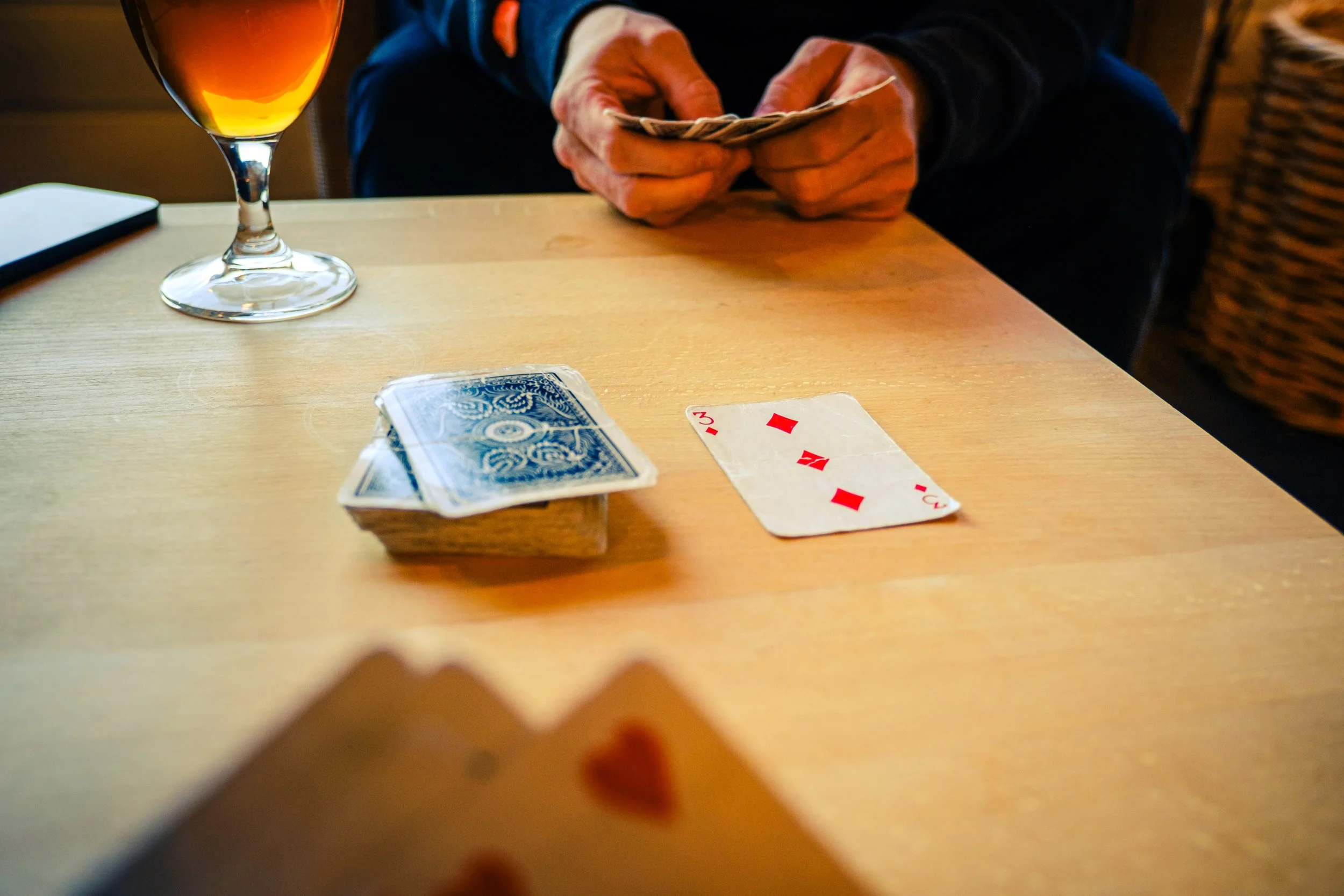 En person holder kort i hendene, med drikke i et glass og et kortspill i bakgrunnen. Kortene ligger på et bord. 