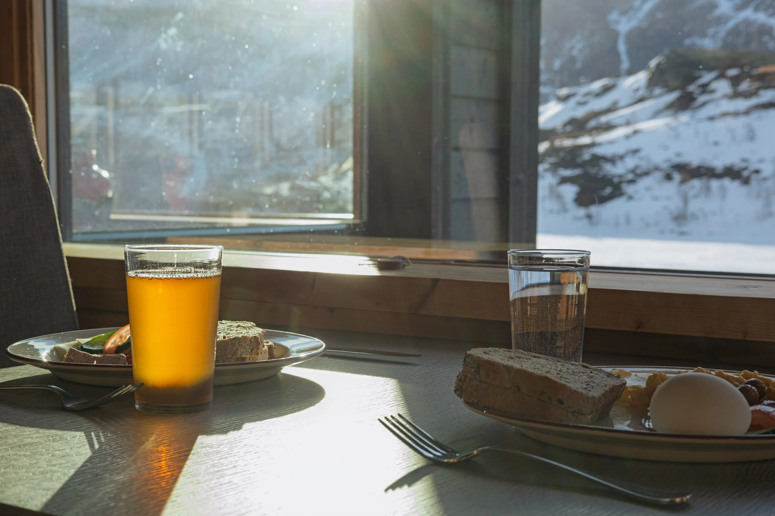 Frokost på et bord med et glass appelsinjuice, et glass vann, et egg, og brødskiver, med et vindu som viser snødekt fjelllandskap i bakgrunnen.