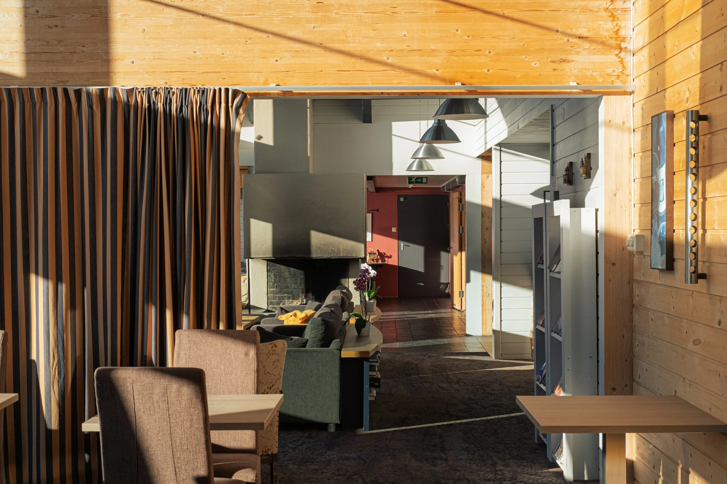 Innendørs bilde av en stue med sofa, blomster, peis og bokhylle,trevegger og dør, med mye dagslys.