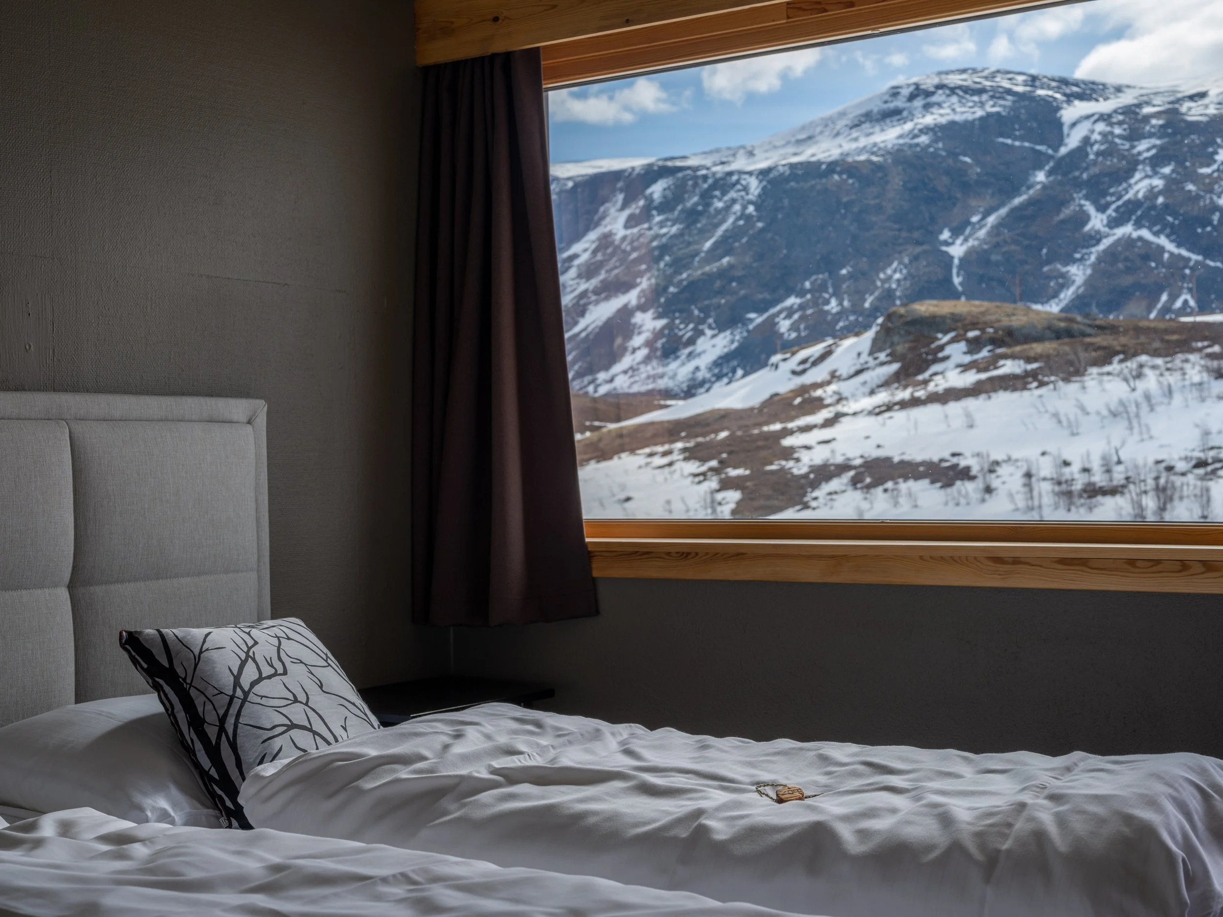 Dobbeltseng med hvit laken og en pute med kvistmønster, ved et vindu som viser snødekte fjell.