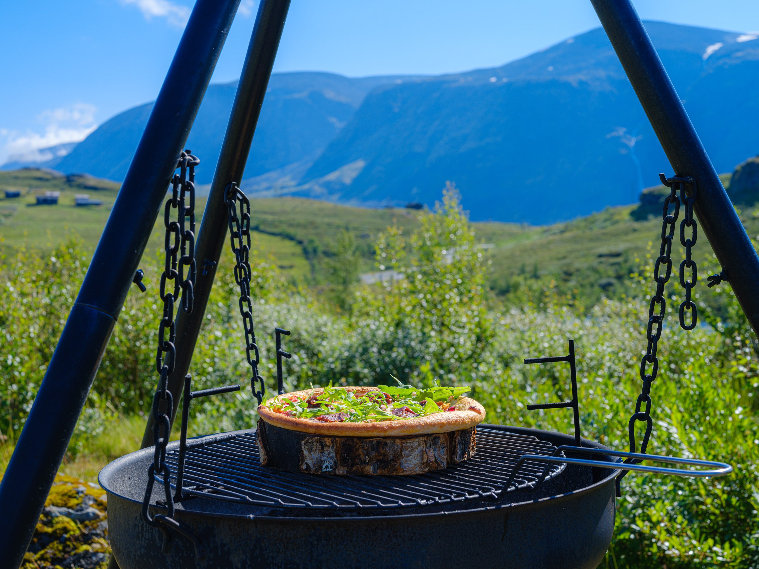 Pizzastein med pizza tilberedt over bål, utendørs med fjell og grønne landskap i bakgrunnen.