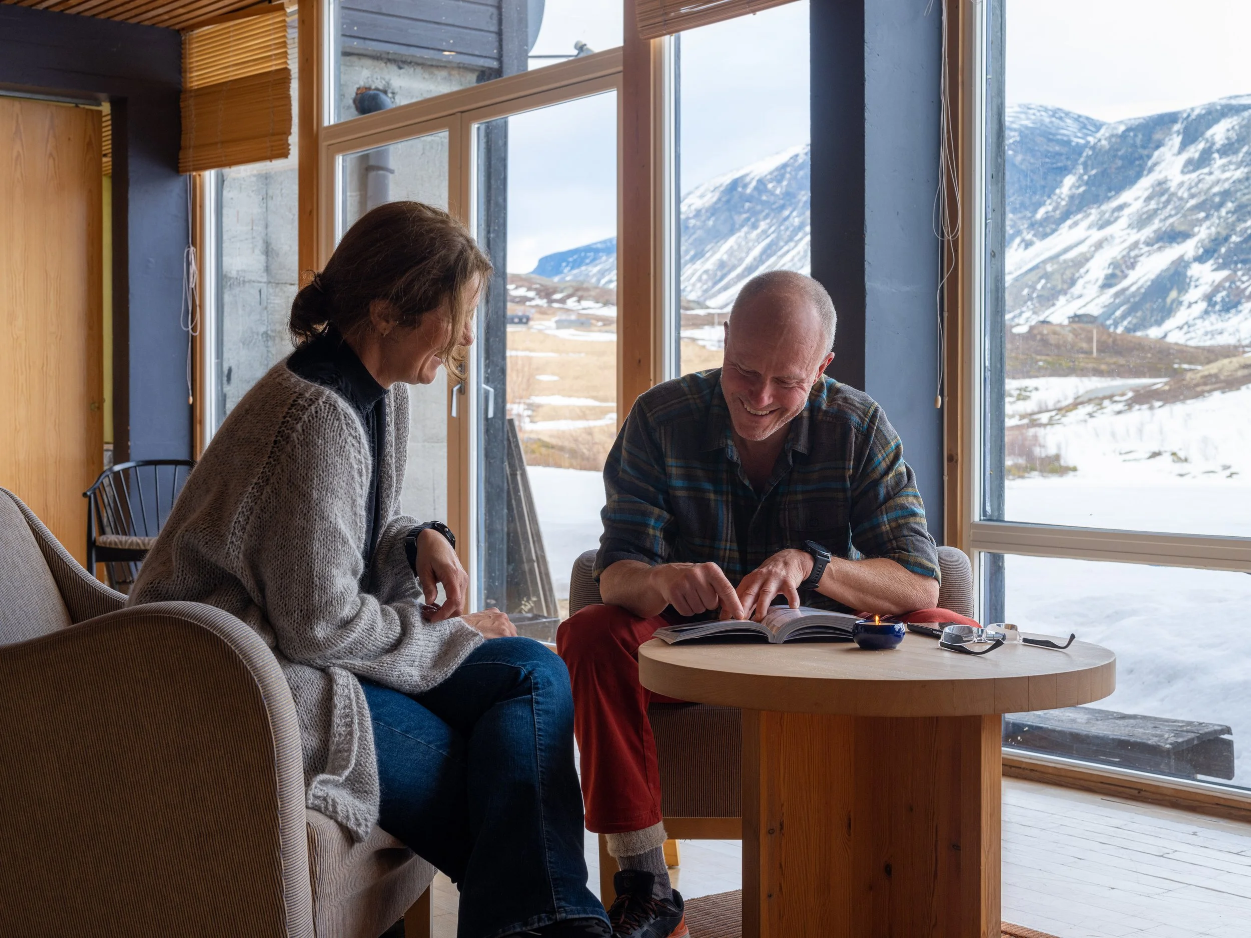 To personer sitter på lenestoler og leser en bok innendørs med snødekte fjell utenfor vinduene.
